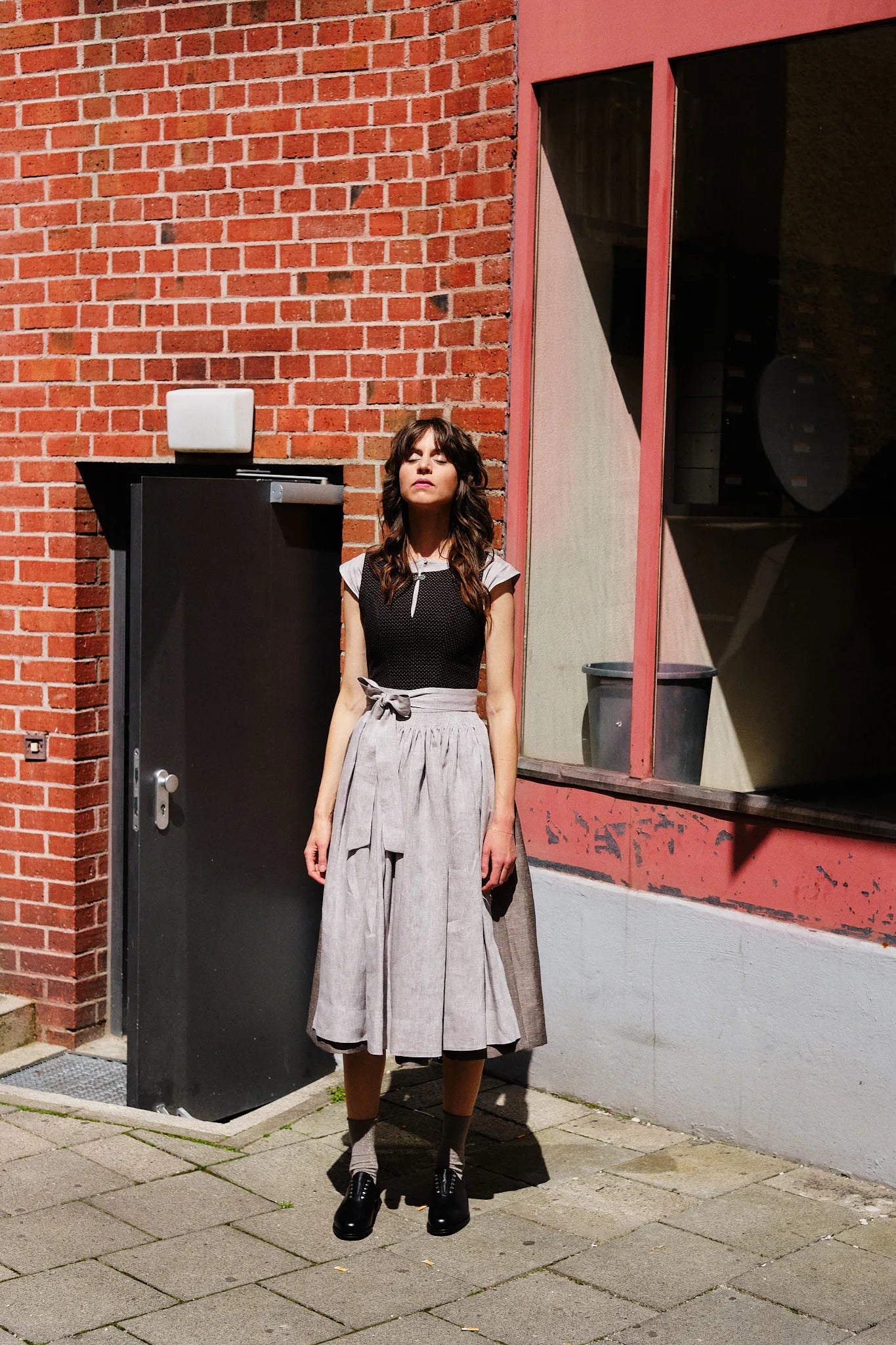 Model trägt Dirndl Marlene in Schwarz-Natur mit Leinenschürze – steht mit geschlossenen Augen der Sonne entgegen vor Backsteinmauer in urbanem Hinterhof, ruhig inszeniert von Zirbenfux