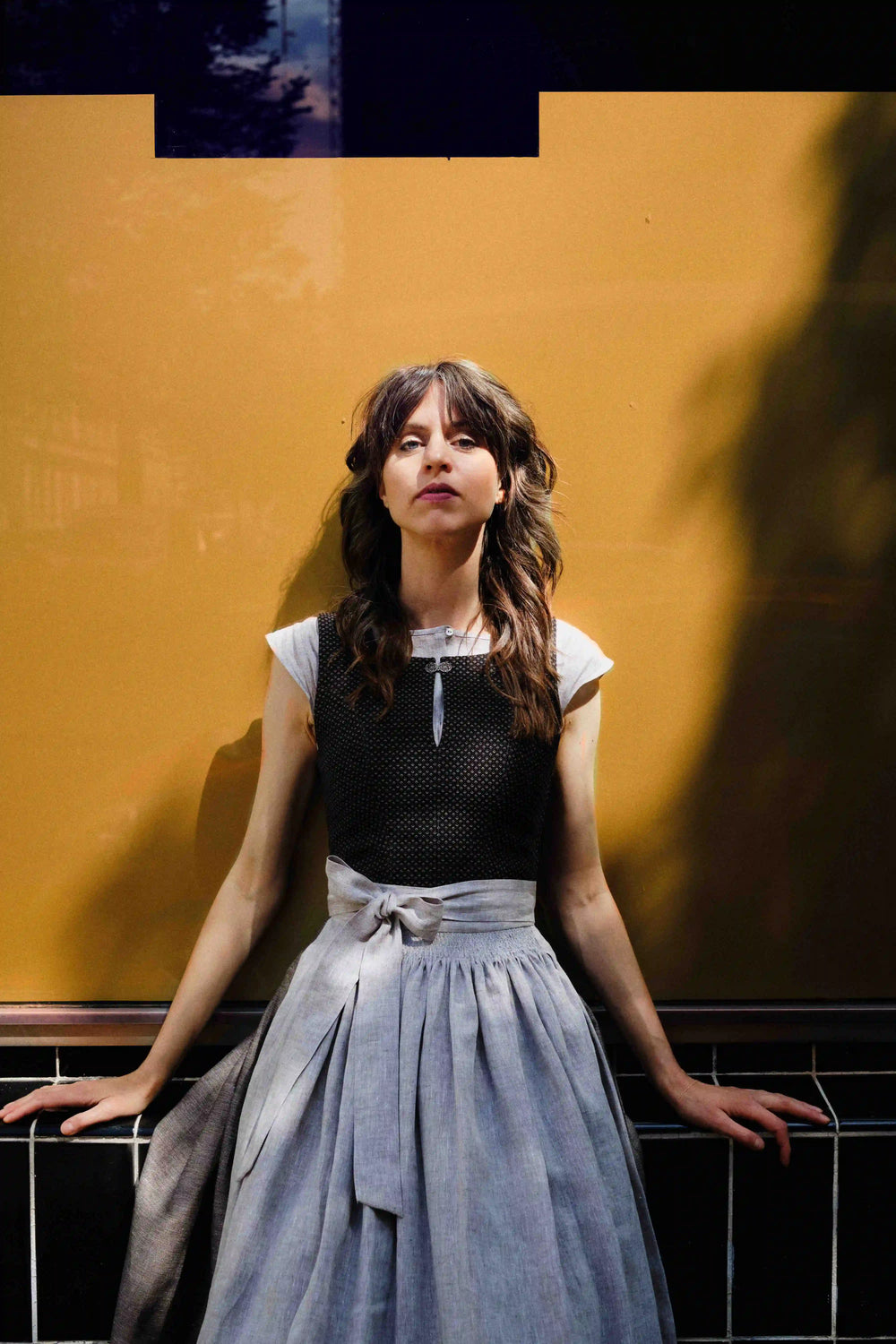 Model trägt Dirndl Marlene in Schwarz-Natur mit Leinenschürze – sitzt vor goldenem Fenster, Hände locker zu beiden Seiten abgelegt, direkter, kühler Blick in die Kamera, Licht und Schattenspiel, stilvoll inszeniert von Zirbenfux