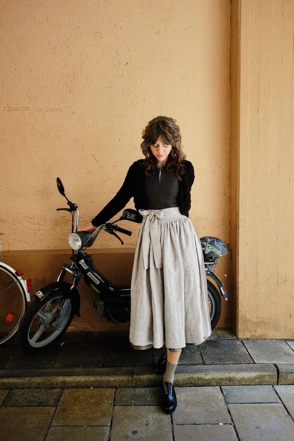 Dirndl Marlene in Schwarz aus Schurwolle-Leinen mit naturfarbener Schürze und Strickjacke, Model in einer Hofeinfahrt vor einem Moped stehend, Arme nach hinten greifend und darauf abgestützt mit Blick nach unten, wertig inszeniert von Zirbenfux.