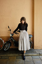 Dirndl Marlene in Schwarz aus Schurwolle-Leinen mit naturfarbener Schürze und Strickjacke, Model in einer Hofeinfahrt vor einem Moped stehend, Arme nach hinten greifend und darauf abgestützt mit Blick nach unten, wertig inszeniert von Zirbenfux.