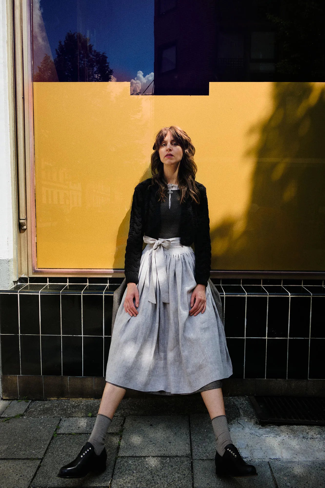 Model trägt Mieder und Rock Marlene in Schwarz-Natur mit Leinenschürze und schwarzer Strickjacke Helene – sitzt mit ausgestreckten Beinen und locker hängenden Armen vor goldenem Fenster, modern interpretiert von Zirbenfux