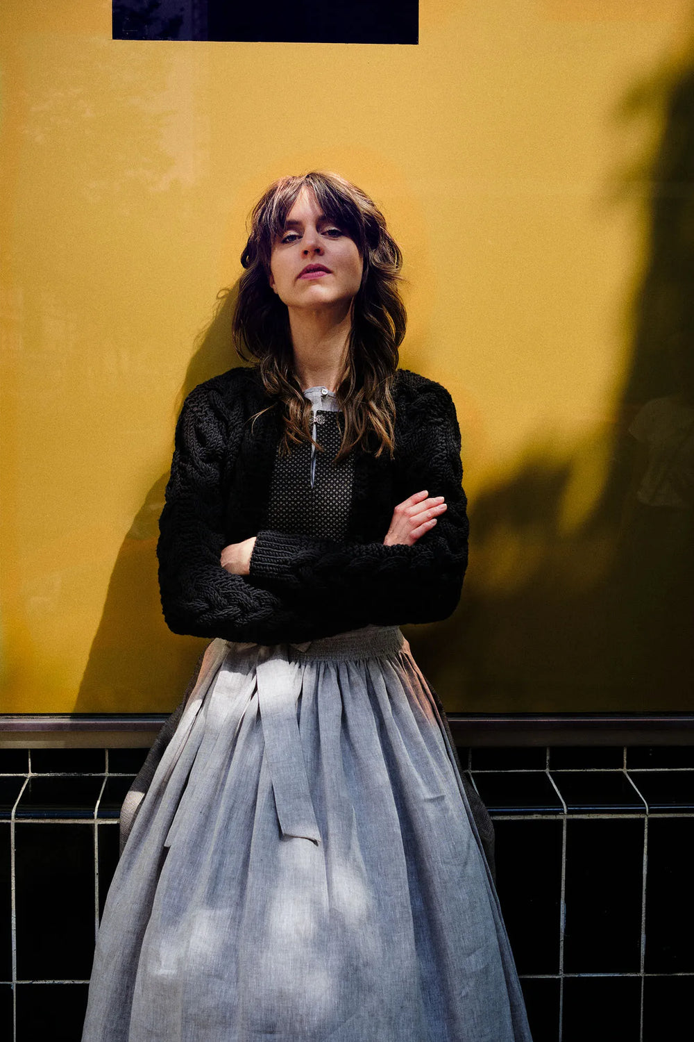 Model trägt Mieder und Rock Marlene in Schwarz-Natur mit Leinenschürze und schwarzer Strickjacke Helene – sitzt mit verschränkten Armen vor goldenem Fenster, direkter Blick in die Kamera, zeitgemäß inszeniert von Zirbenfux