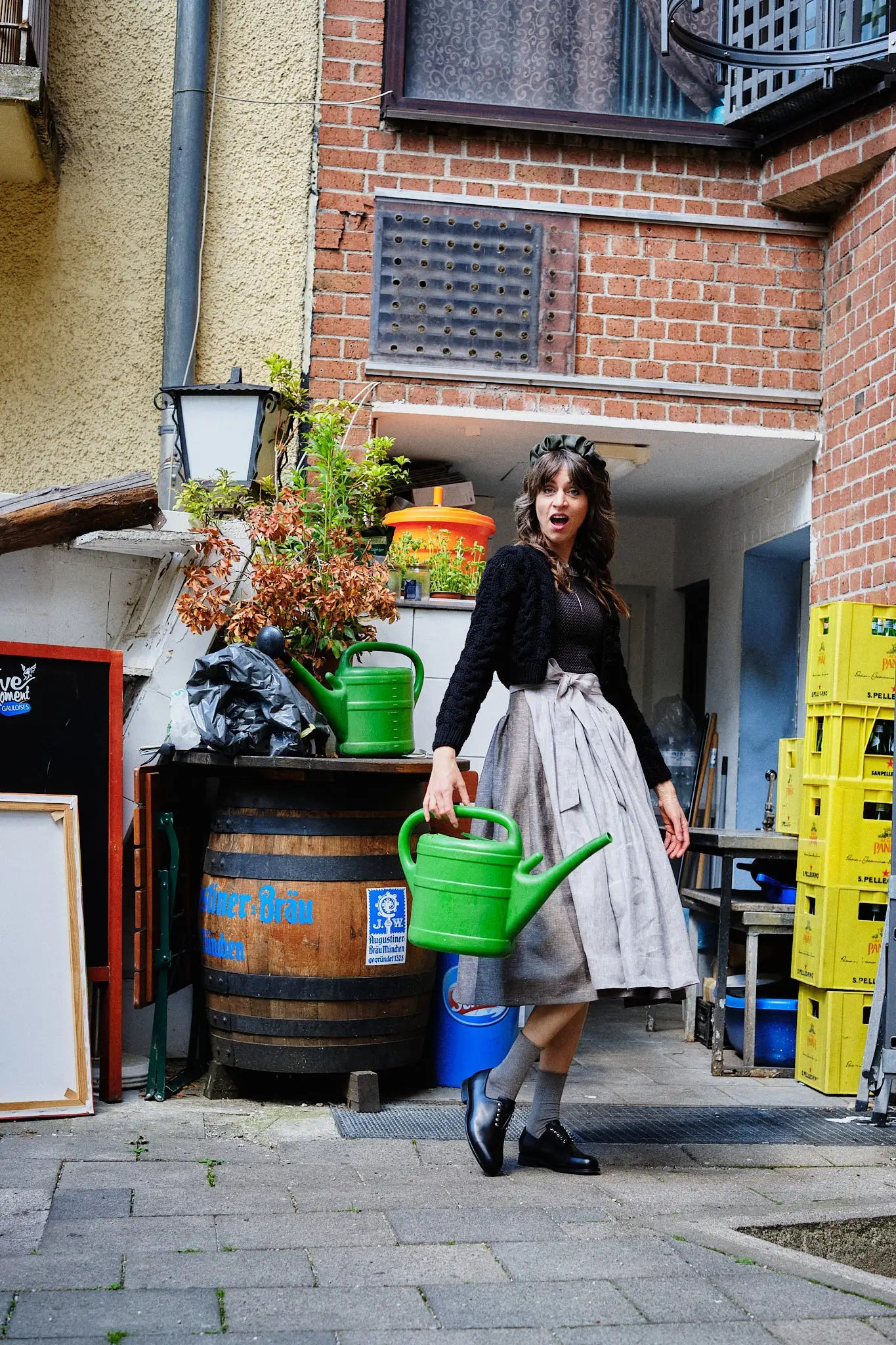 Dirndl Marlene in Schwarz aus Schurwolle-Leinen mit naturfarbener Schürze und Strickjacke, Model im Hinterhof am Hintereingang eines Restaurants zwischen verschiedenen Gegenständen stehend, große grüne Gießkanne in der Hand und scherzhafte Pose, spielerisch inszeniert von Zirbenfux.