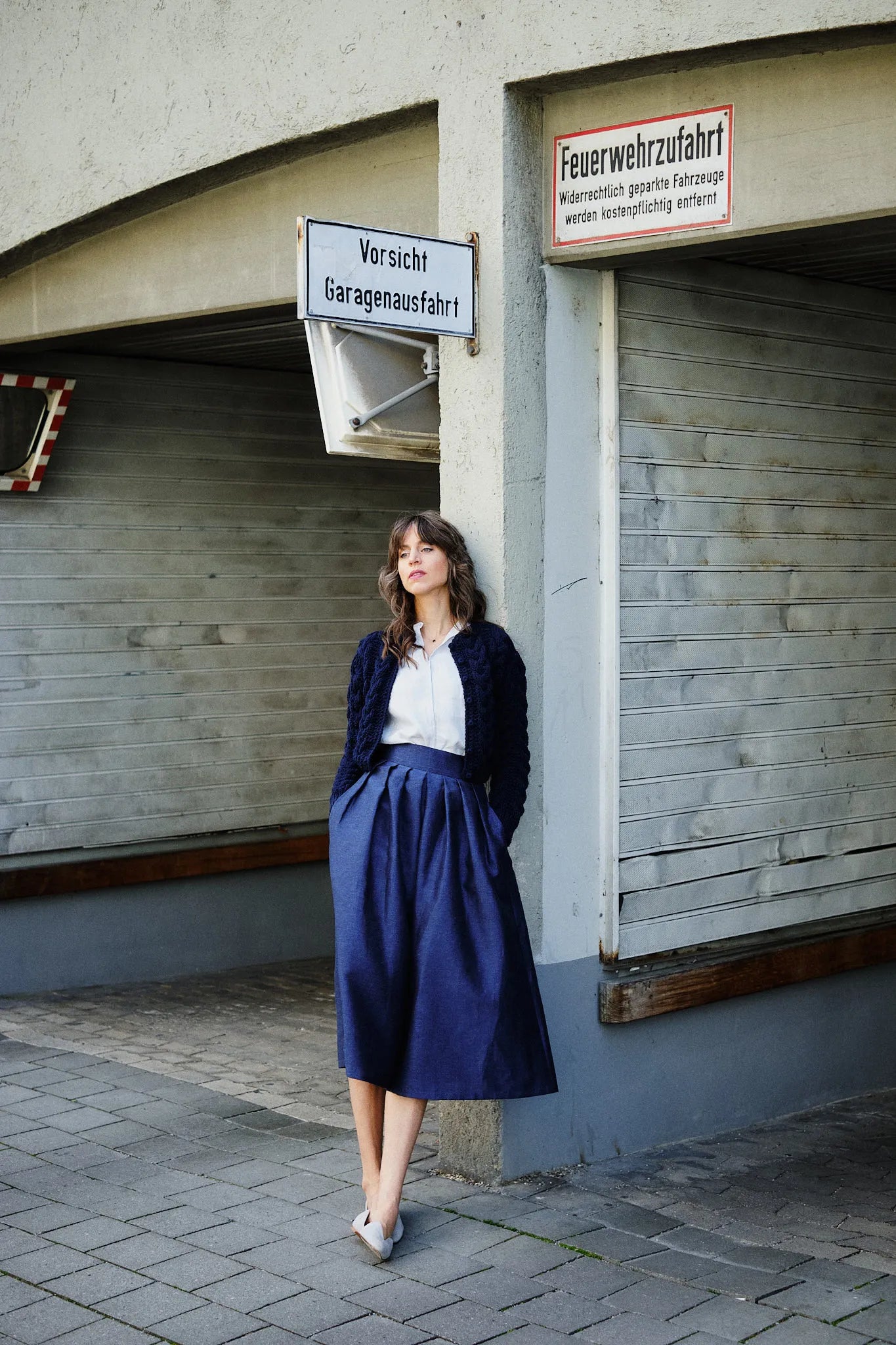 Hosenrock Audrey mit Bluse Romy und Strickjacke im Set in Blau aus Leinen, Model an eine Garageneinfahrt angelehnt im urbanen Setting mit unbestimmtem Blick, zeitgemäß interpretiert von Zirbenfux.