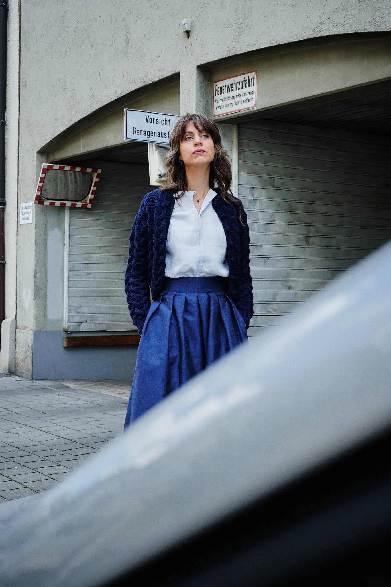 Hosenrock Audrey mit Bluse Romy und Strickjacke im Set in Blau aus Leinen, Model stehend in einer urbanen Garageneinfahrt mit Blick in die Ferne, Auto im Anschnitt im Vordergrund, ruhig interpretiert von Zirbenfux.