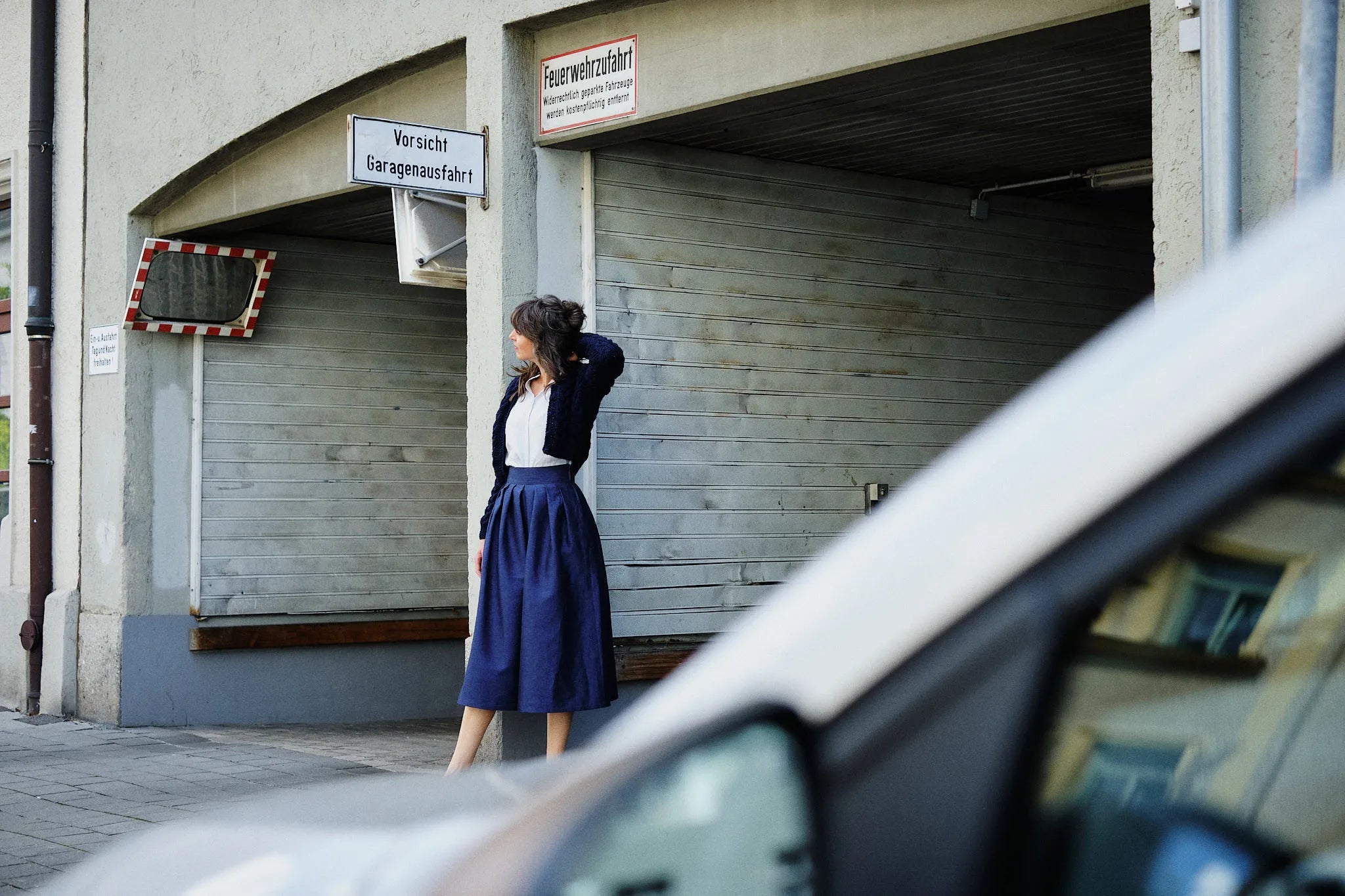 Hosenrock Audrey mit Bluse Romy und Strickjacke im Set in Blau aus Leinen, Model stehend an einer urbanen Garageneinfahrt mit abgewandtem Blick, Auto im Anschnitt im Vordergrund, klar inszeniert von Zirbenfux.