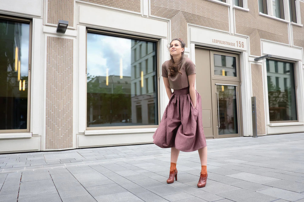 Hosenrock Audrey mit Fuxerl TILDA im Set in Malve aus Leinen, Model stehend vor moderner urbaner Häuserfassade, windige Szene mit Hosenrock in Bewegung, markant inszeniert von Zirbenfux.