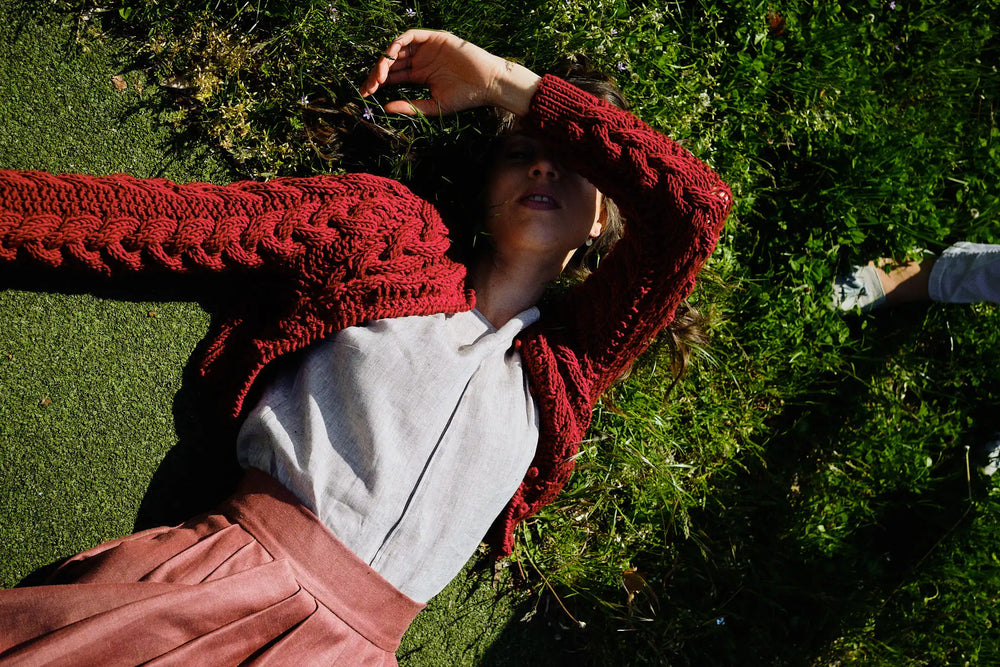 Strickjacke Helene in Kirsche aus Merinowolle mit Bluse TILDA aus Leinen in Natur, Model auf dem Rückend liegend in einer Wiese und sich mit dem Ellbogen selbst Schatten spendend, ruhig inszeniert von Zirbenfux.