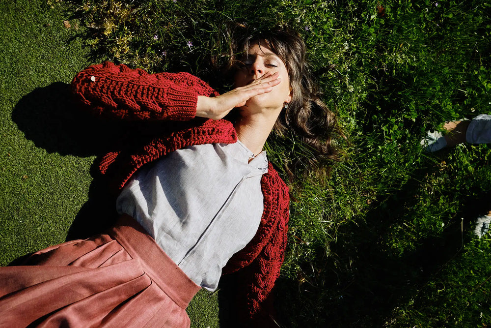 Model mit geschlossenen Augen im Gras – trägt Strickjacke Helene in Kirschrot, naturfarbene Leinenbluse Tilda und Rock in pinker Beere, handgefertigt in München von Zirbenfux