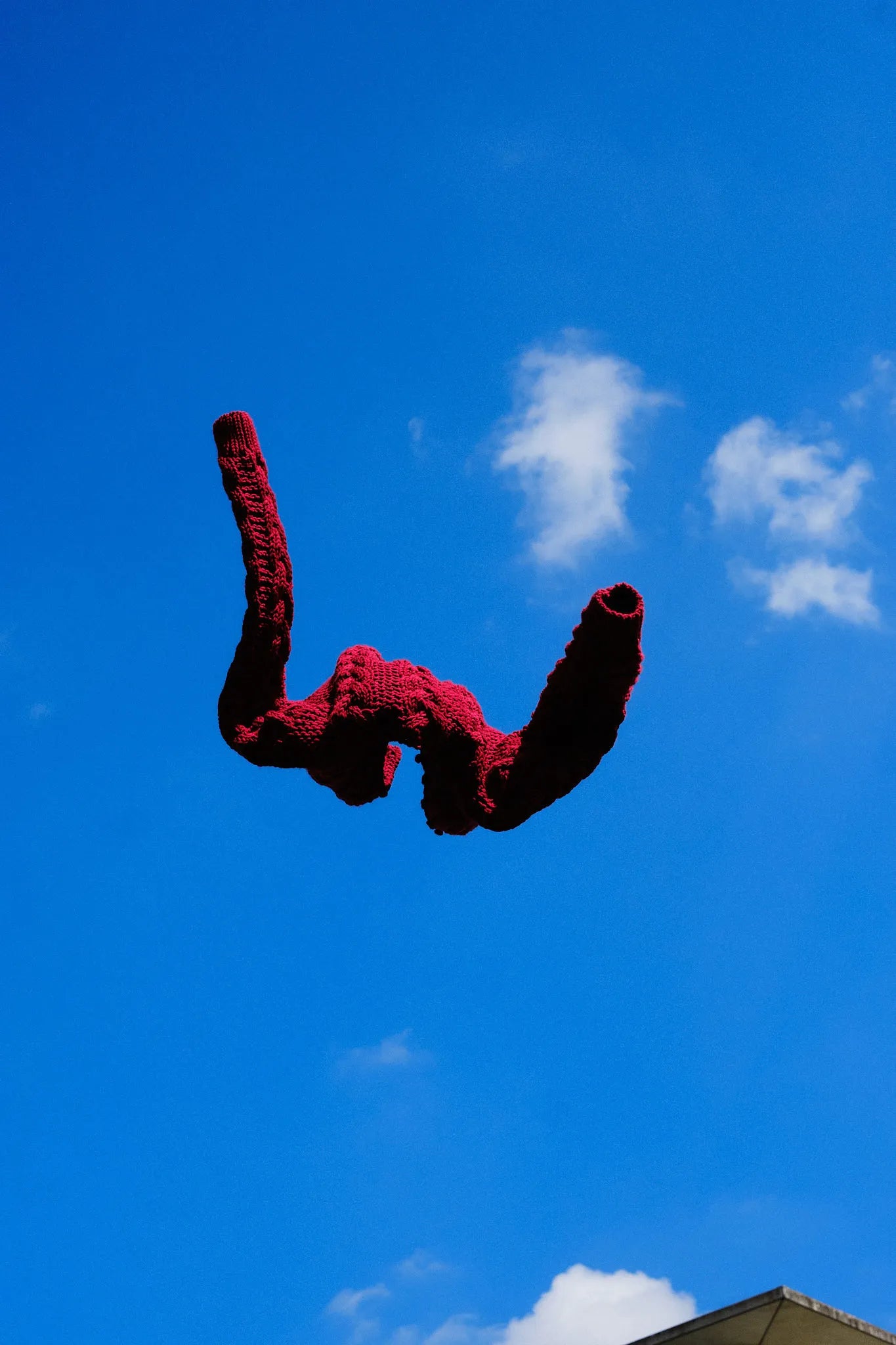 Strickjacke Helene in Kirsche aus Merinowolle, frei in der Luft fliegend vor strahlend blauem Himmel mit weißen Wolken, dynamisch inszeniert von Zirbenfux.