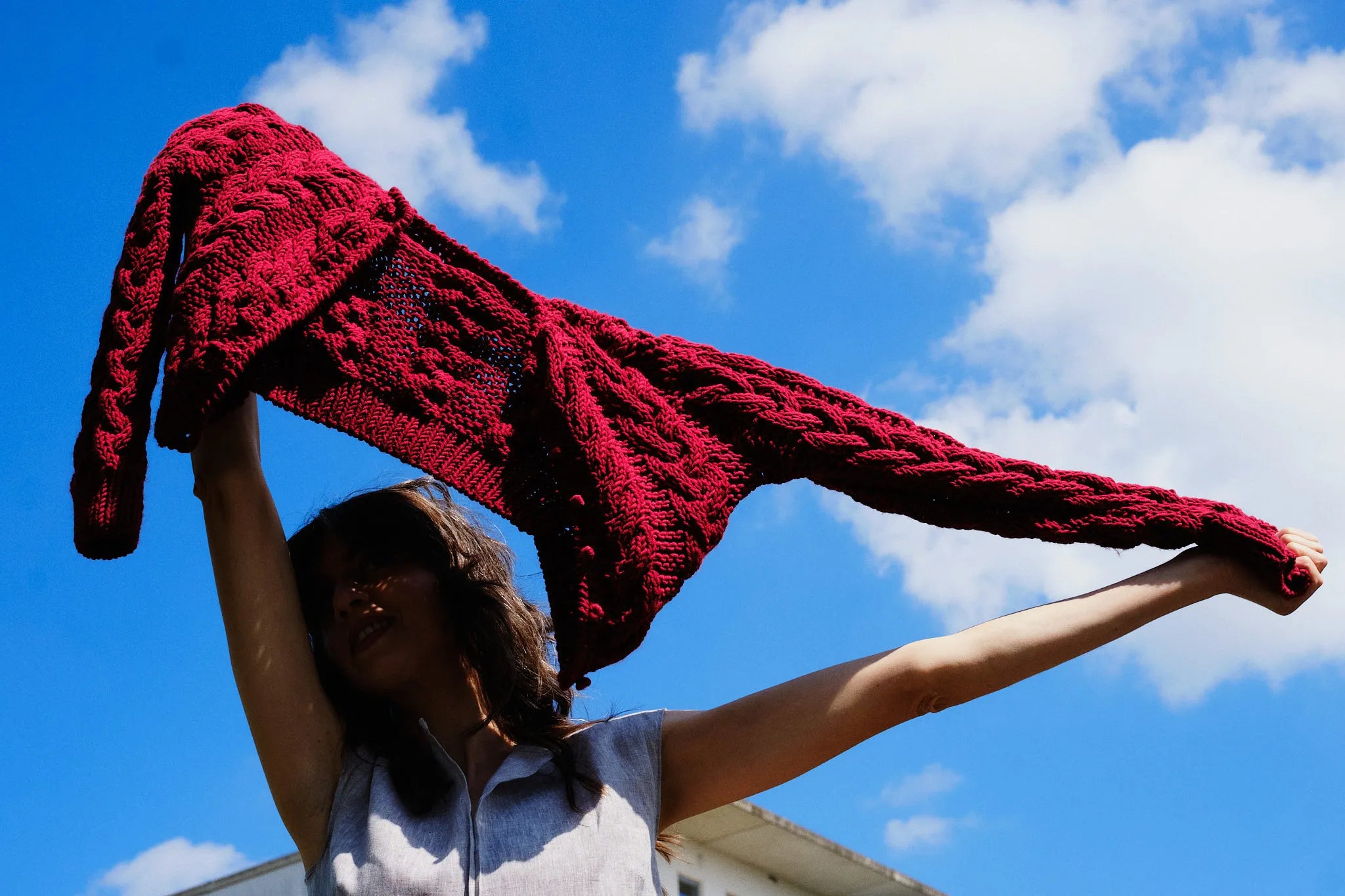 Strickjacke Helene aus Merinowolle in Kirschrot – vom Model über den Kopf gespannt, im Gegenlicht vor blauem Himmel mit Wolken, urbane Tracht von Zirbenfux