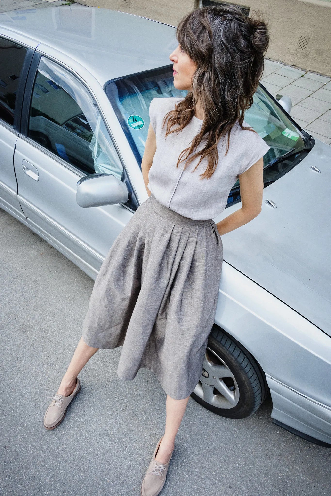 Model trägt Trachtenrock Marlene aus goldbrauner Leinen-Schurwolle-Mischung und Bluse Tilda aus naturfarbenem Bio-Leinen – lehnt lässig an silbernem Auto in urbanem Umfeld, Blick zur gegenüberliegenden Seite, zeitgemäß interpretiert von Zirbenfux