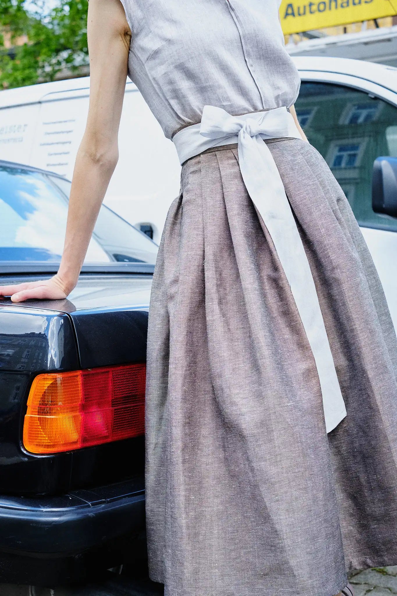 Nahaufnahme von Trachtenrock Marlene in goldbraun, Schürzenband und Leinenbluse Tilda in Natur – Model stützt sich auf Kofferraumklappe in einem urbanen Setting, Bildausschnitt zeigt nur den Körper, moderne Tracht von Zirbenfux