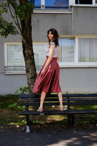 Trachtenrock Marlene in Pinke Beere mit Bluse TILDA im Set aus Leinen in Natur, Model gerade auf einer Bank stehend mit dem Rücken zum Betrachter und über die Schulter in die Kamera blickend, ruhig inszeniert von Zirbenfux.