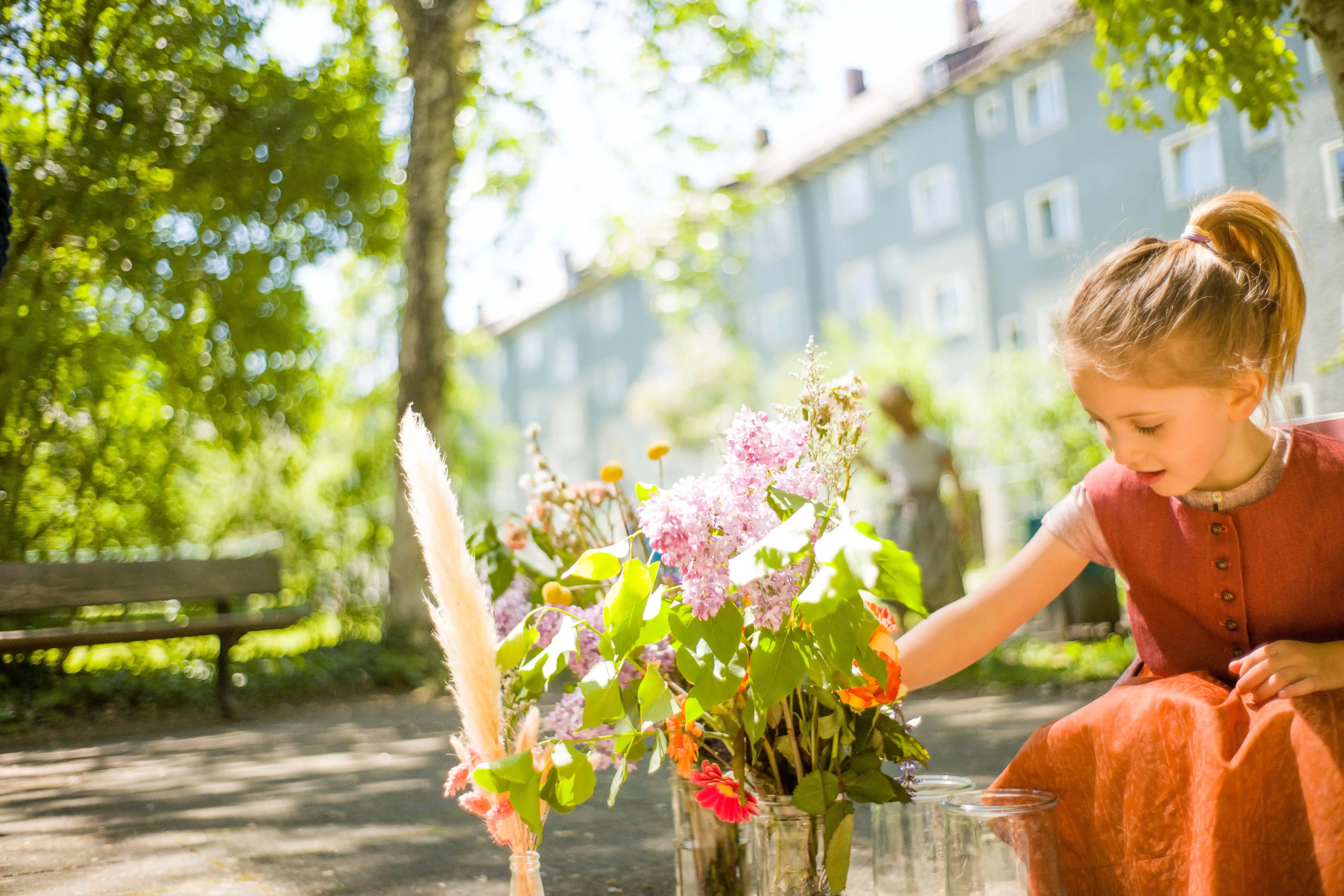 Stimmungsbild von Zirbenfüxlein mit pinker Beere Kinderdirndl, Kind in der Hocke einen Blumenstrauß in grüner Hinterhofszene arrangierend, ruhig interpretiert von Zirbenfüxlein.