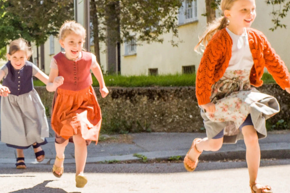 Zirbenfüxlein Kinderdirndl in pinker Beere, Saphir und Tintenblau, drei Kinder rennen lachend über eine sonnige Straße in urbaner Szene, dynamisch gestaltet von Zirbenfüxlein.