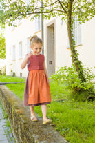 Kinderdirndl in pinker Beere mit terrakottafarbener Schürze – balancierend auf einem Mäuerchen vor urbaner Häuserfront, modern inszeniert von Zirbenfüxlein