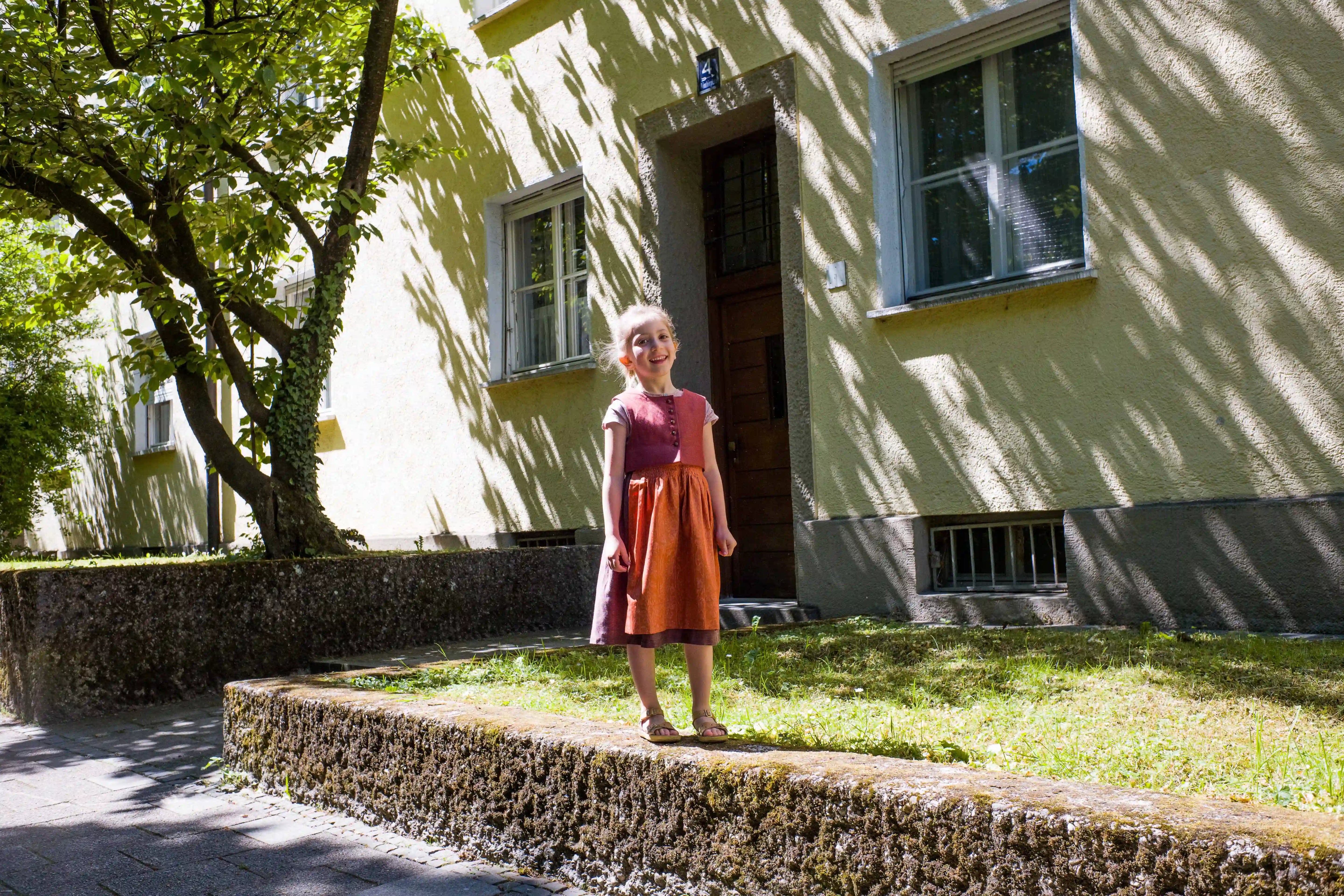 Kinderdirndl in pinker Beere mit terrakottafarbener Schürze – stehend auf einem Mäuerchen vor urbaner Häuserfront, markantes Licht- und Schattenspiel, modern inszeniert von Zirbenfüxlein