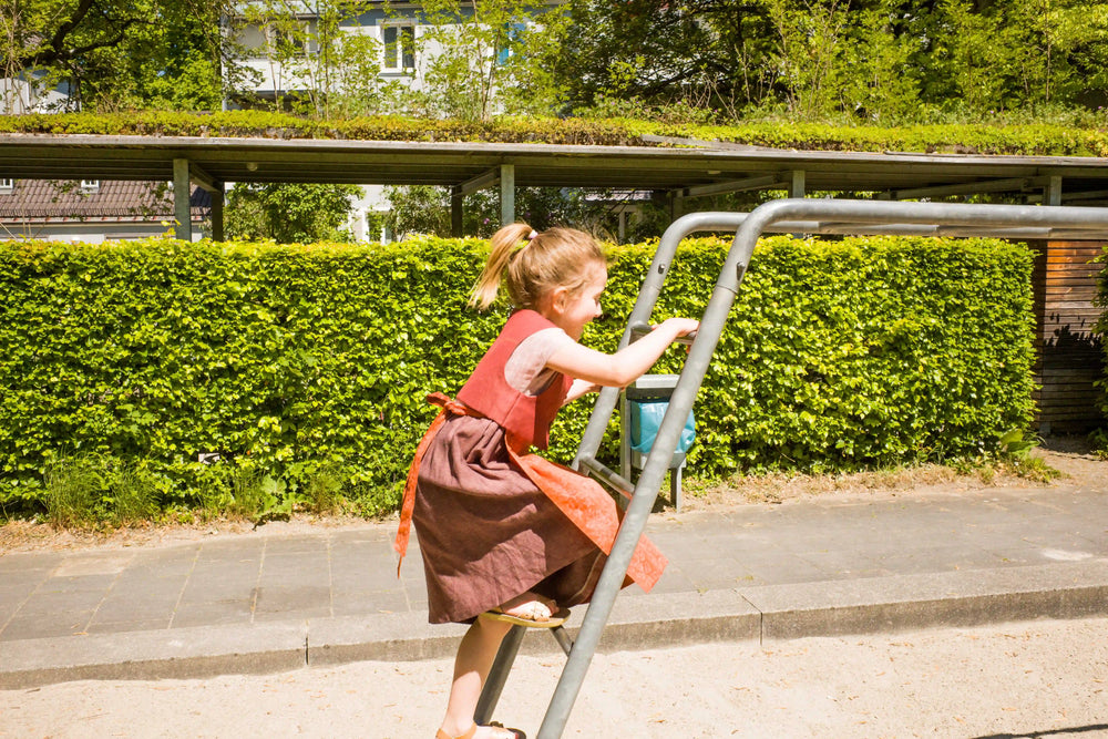 Kinderdirndl in pinker Beere mit terrakottafarbener Schürze – auf eine Spielplatz-Leiter kletternd, aktiv und alltagsnah inszeniert von Zirbenfüxlein