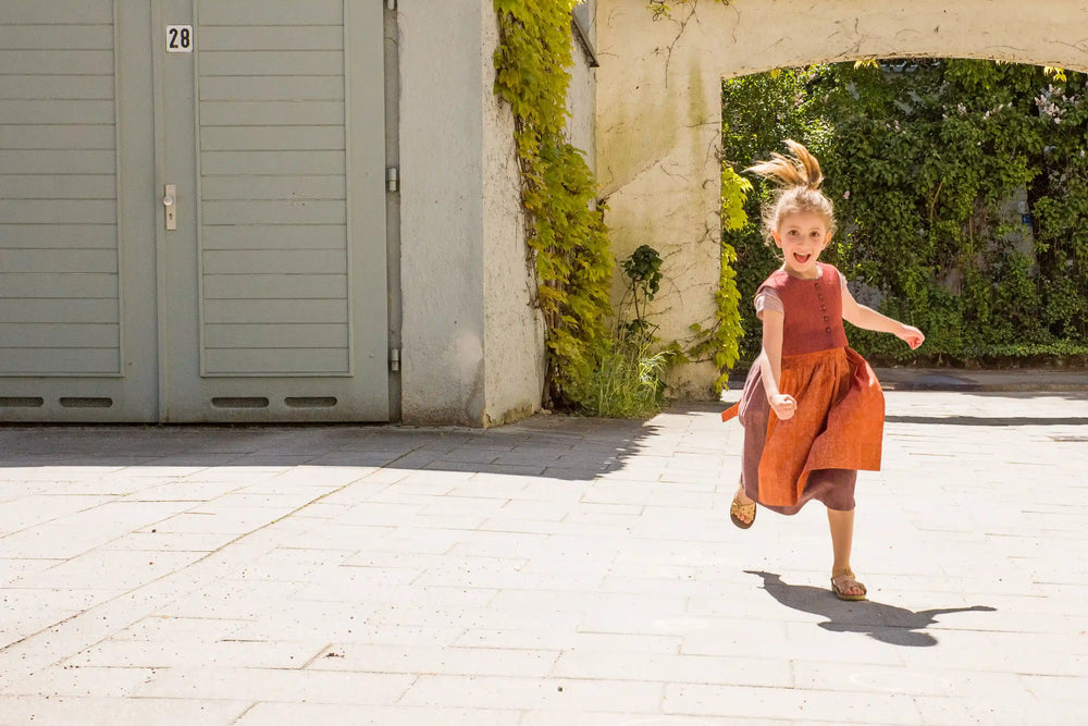 Kinderdirndl in pinker Beere mit terrakottafarbener Schürze – Kind rennt nach vorne in einem sonnigen Hinterhof, energiegeladen inszeniert von Zirbenfüxlein