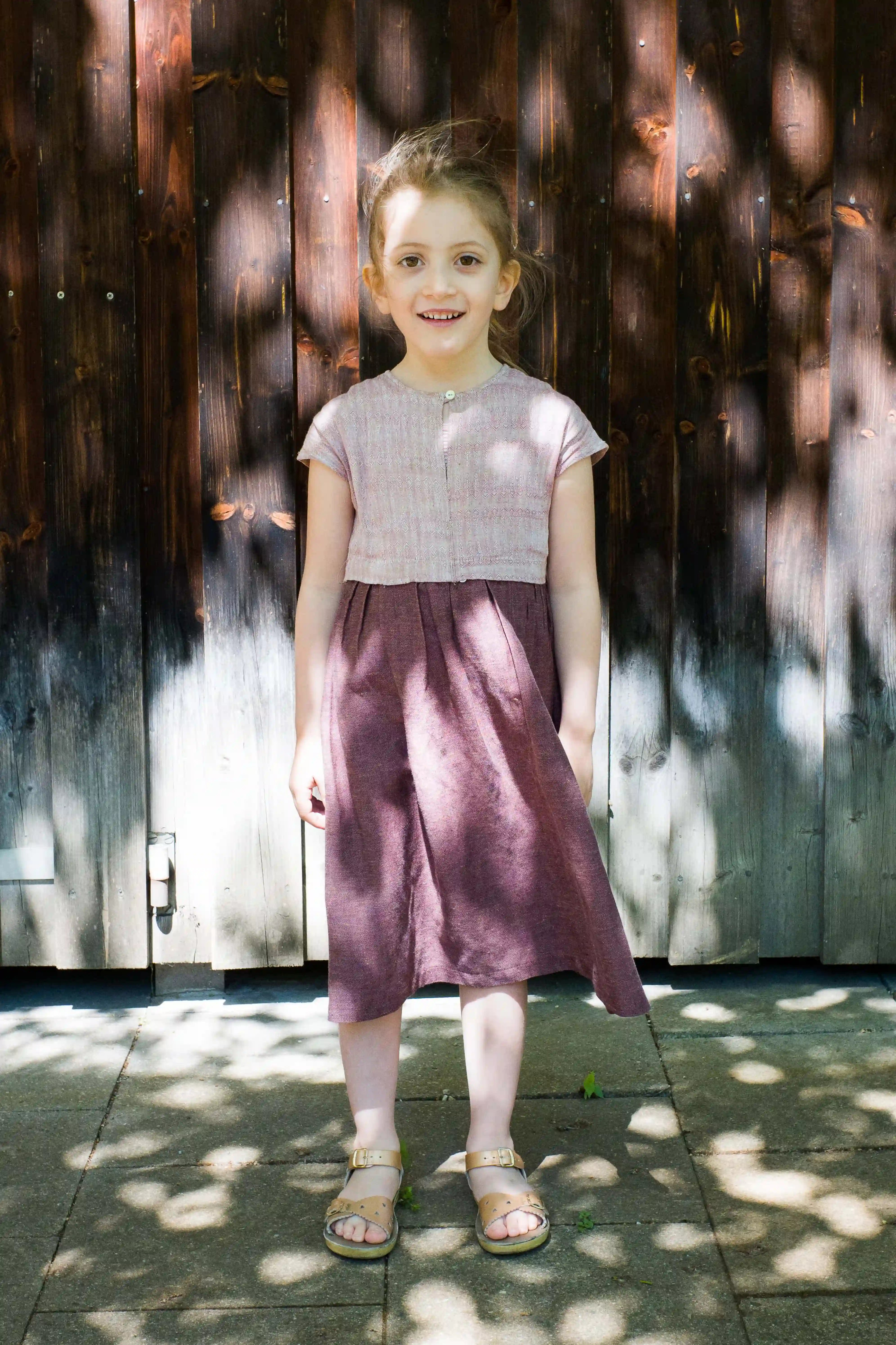 Kinderdirndl in Rosenholz – Frontansicht vor Altholzwand auf Asphalt, mit kontrastreichem Schattenspiel, ruhig inszeniert von Zirbenfüxlein