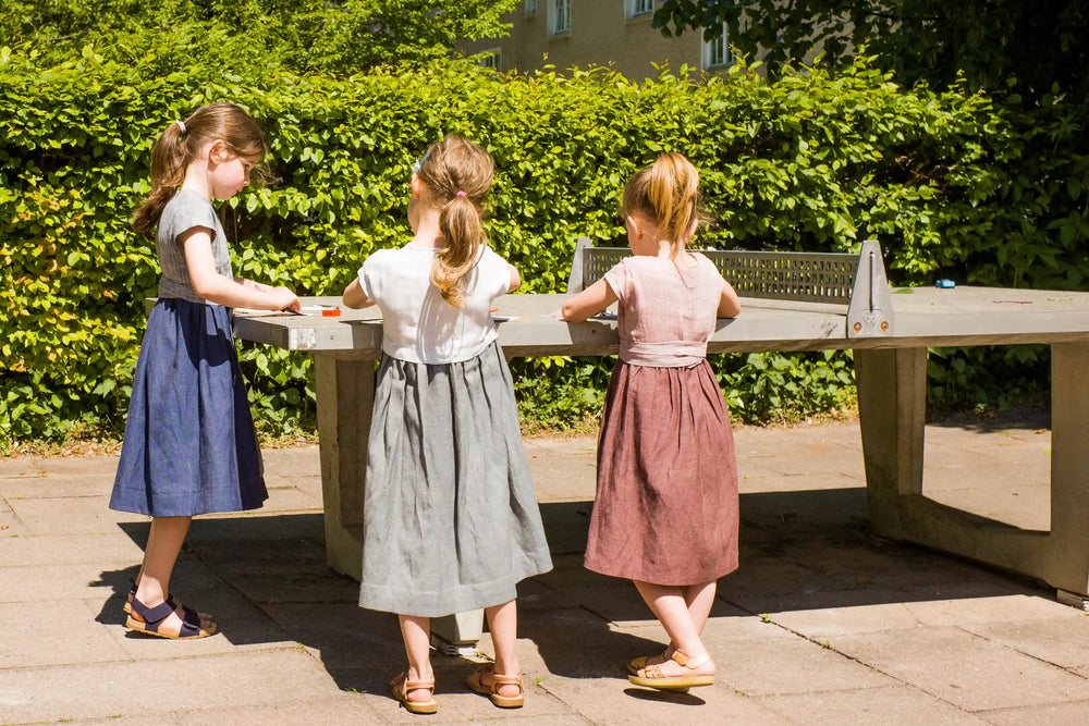 Zirbenfüxlein Kinderdirndl in Rosenholz, Saphir und Tintenblau, drei Kinder an einer Tischtennisplatte im urbanen Hinterhof stehend und zeichnend dargestellt, ruhig umgesetzt von Zirbenfüxlein.