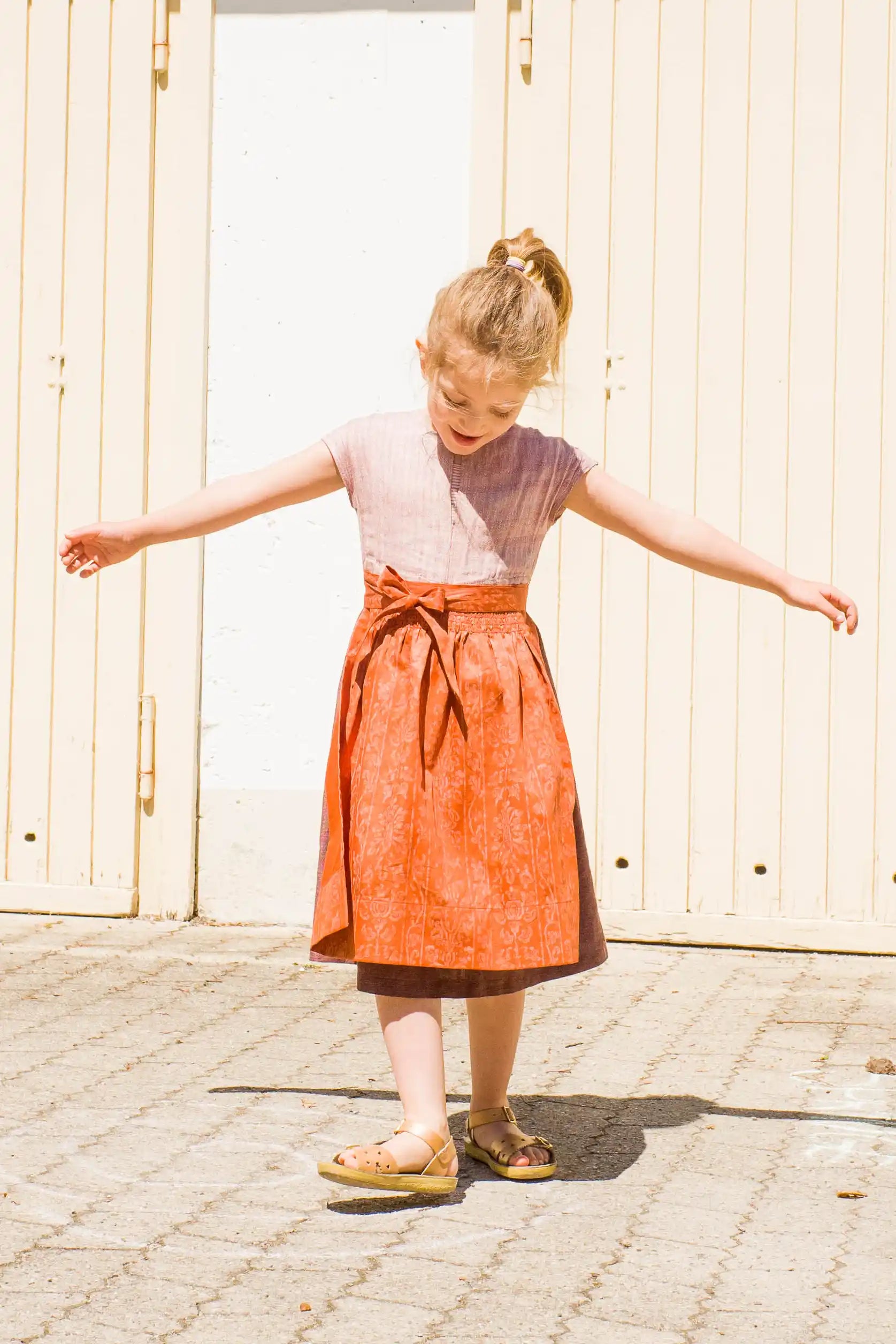Kinderdirndl in Rosenholz mit terrakottafarbener Schürze – Frontansicht beim Drehen vor Garagentor, Blick nach unten, Arme seitlich ausgestreckt, sonnige Szene auf Asphalt, lebendig interpretiert von Zirbenfüxlein