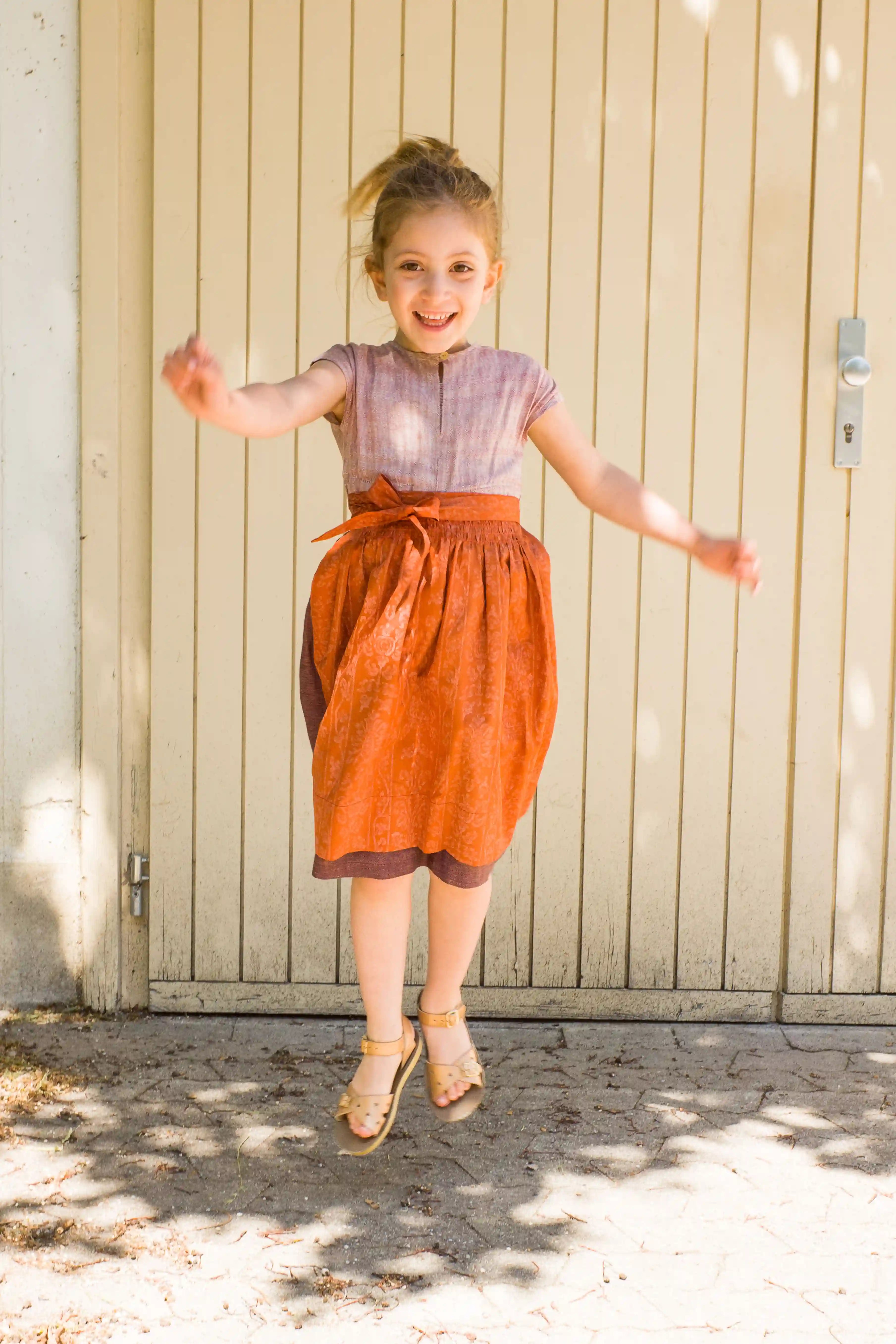 Kinderdirndl in Rosenholz mit terrakottafarbener Schürze – springend und lachend vor einem Garagentor, lebendige Szene mit Schattenspiel, spielerisch inszeniert von Zirbenfüxlein