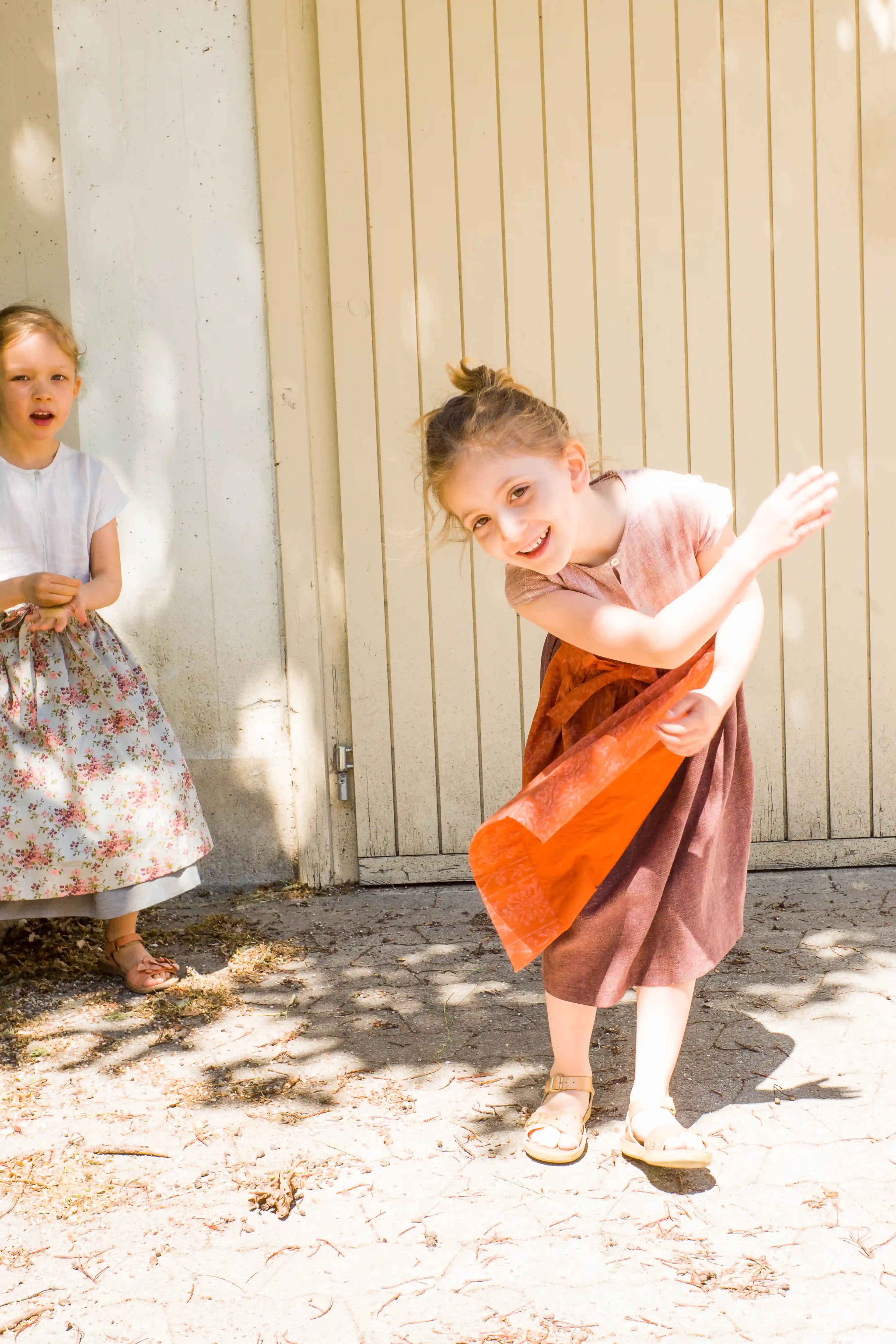 Kinderdirndl in Rosenholz mit terrakottafarbener Schürze – springendes Kind vor einem Garagentor, ein weiteres Kind im Hintergrund, lebendige Situation, natürlich inszeniert von Zirbenfüxlein
