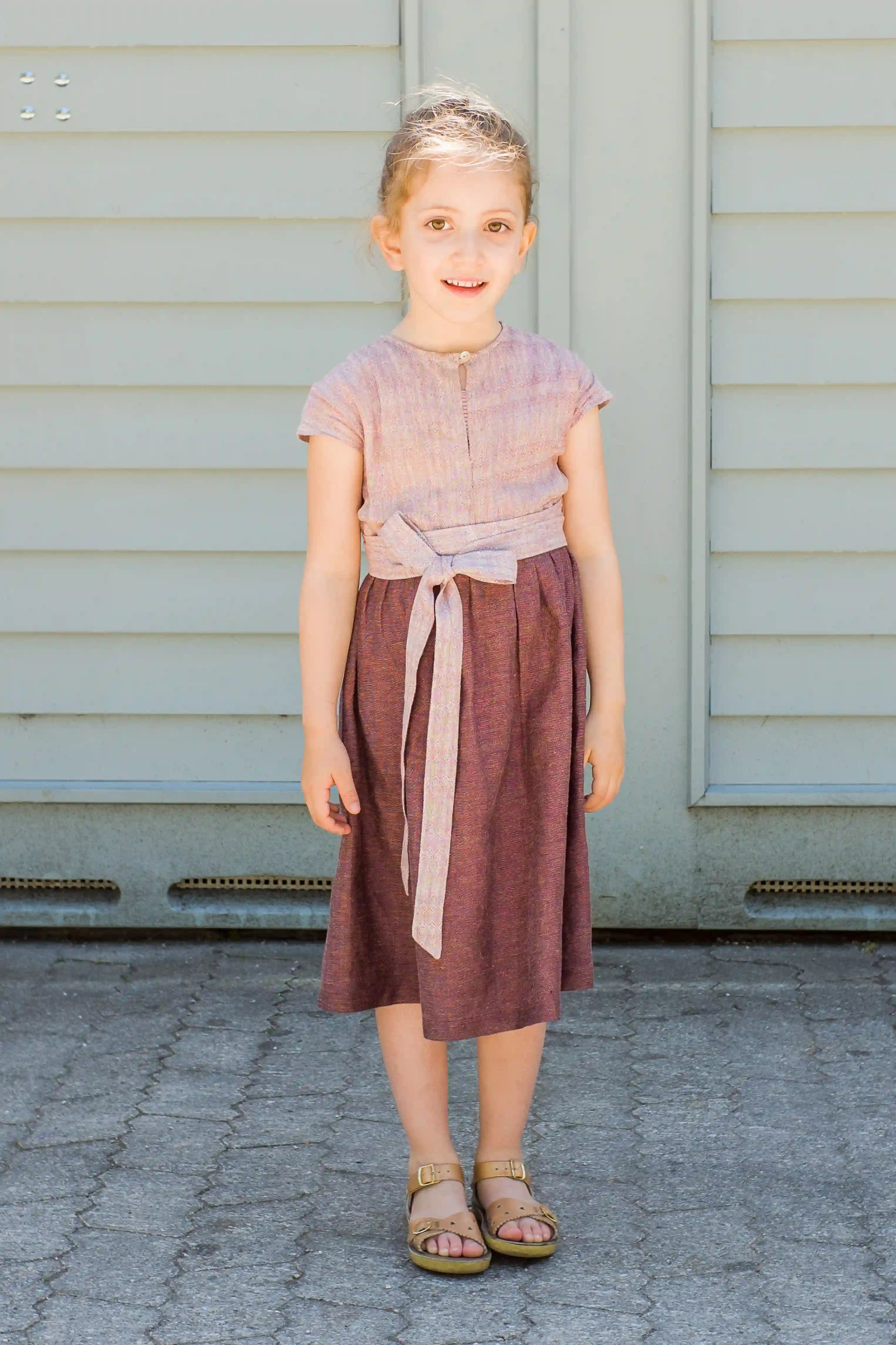 Kinderdirndl in Rosenholz mit Schürzenband – frontal und ruhig stehend vor einem Garagentor, klar inszeniert von Zirbenfüxlein