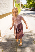 Kinderdirndl in Rosenholz mit Schürzenband – Kind läuft auf dem Gehweg entlang einer Straße im urbanen Setting, fröhlich und modern inszeniert von Zirbenfüxlein