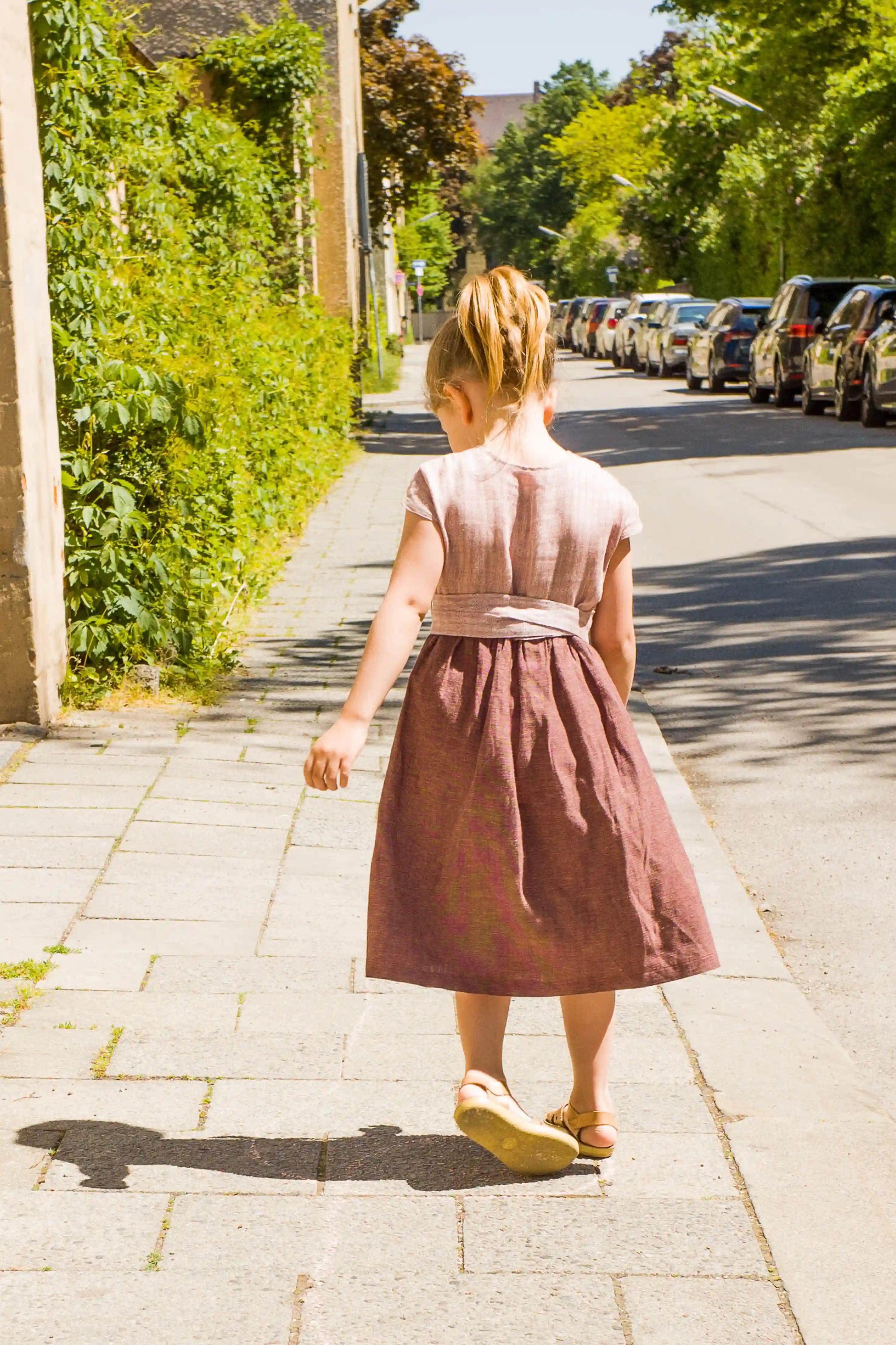 Kinderdirndl in Rosenholz mit Schürzenband – Kind dreht sich mit Blick nach unten auf einem Gehweg an einer Straße, Rückansicht, lebendig inszeniert von Zirbenfüxlein