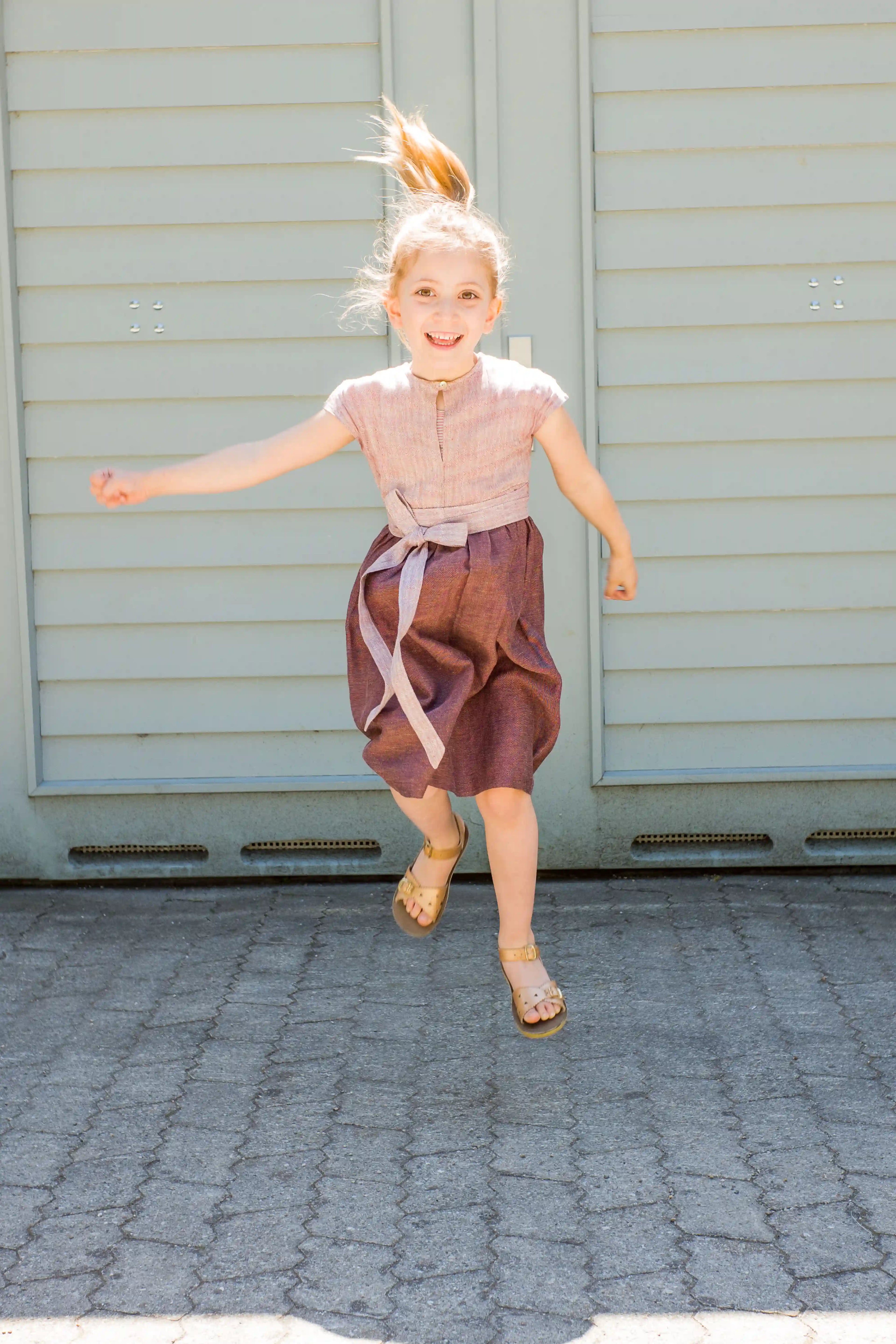 Kinderdirndl in Rosenholz mit Schürzenband – fröhlich nach oben springend vor einem Garagentor im Sonnenschein, dynamisch und natürlich inszeniert von Zirbenfüxlein