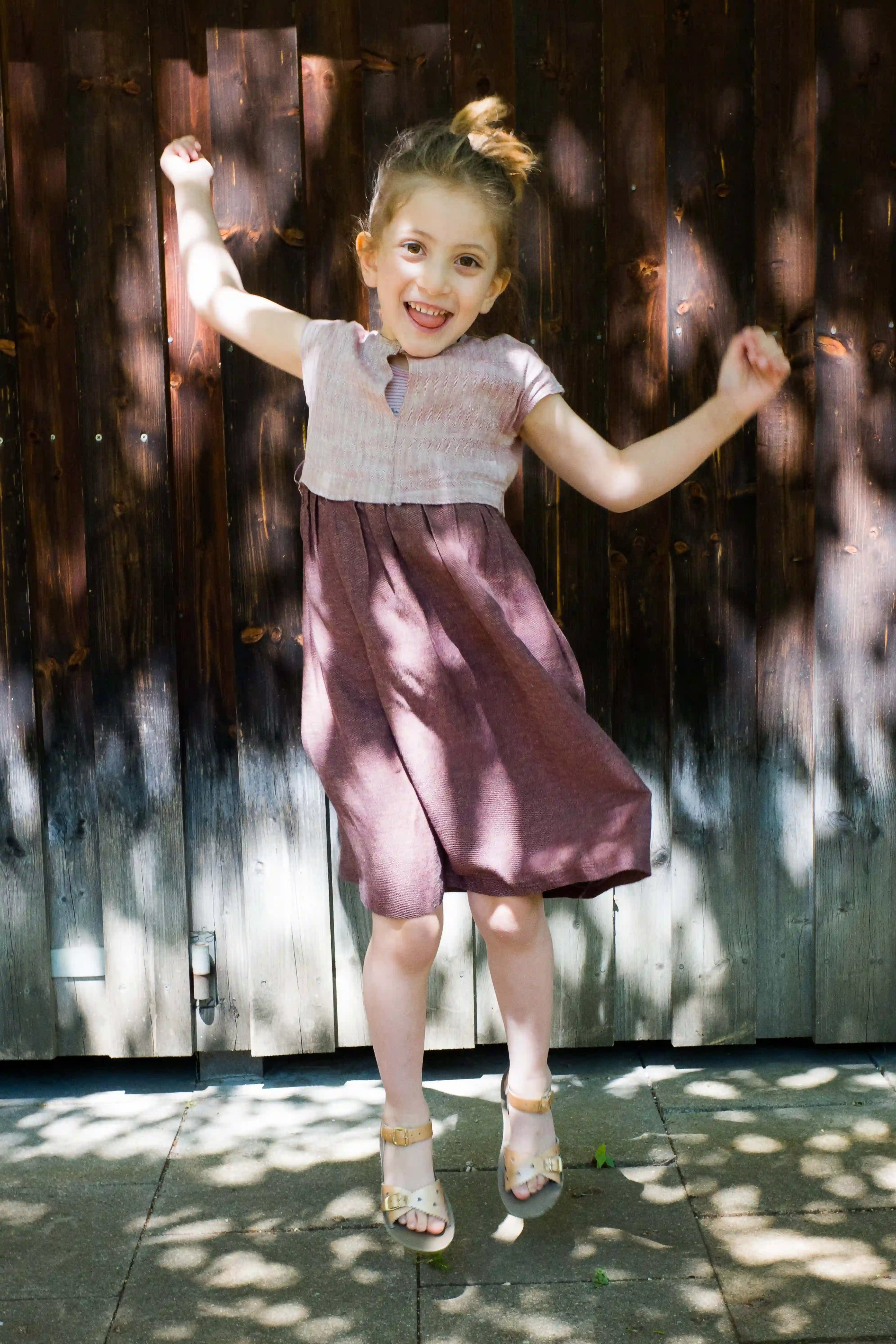 Kinderdirndl in Rosenholz – fröhlich springend vor Altholzwand auf Asphalt, mit kontrastreichem Schattenspiel, spielerisch inszeniert von Zirbenfüxlein