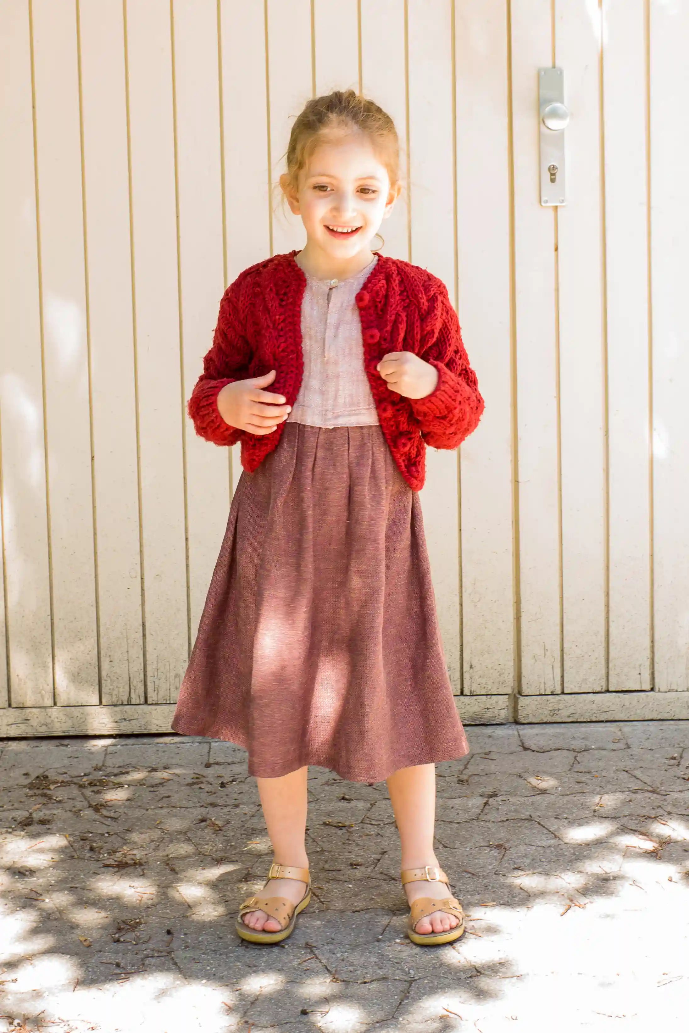 Kinderdirndl in Rosenholz mit kirschroter Strickjacke – locker und frontal stehend vor einem Garagentor, stilvoll inszeniert von Zirbenfüxlein