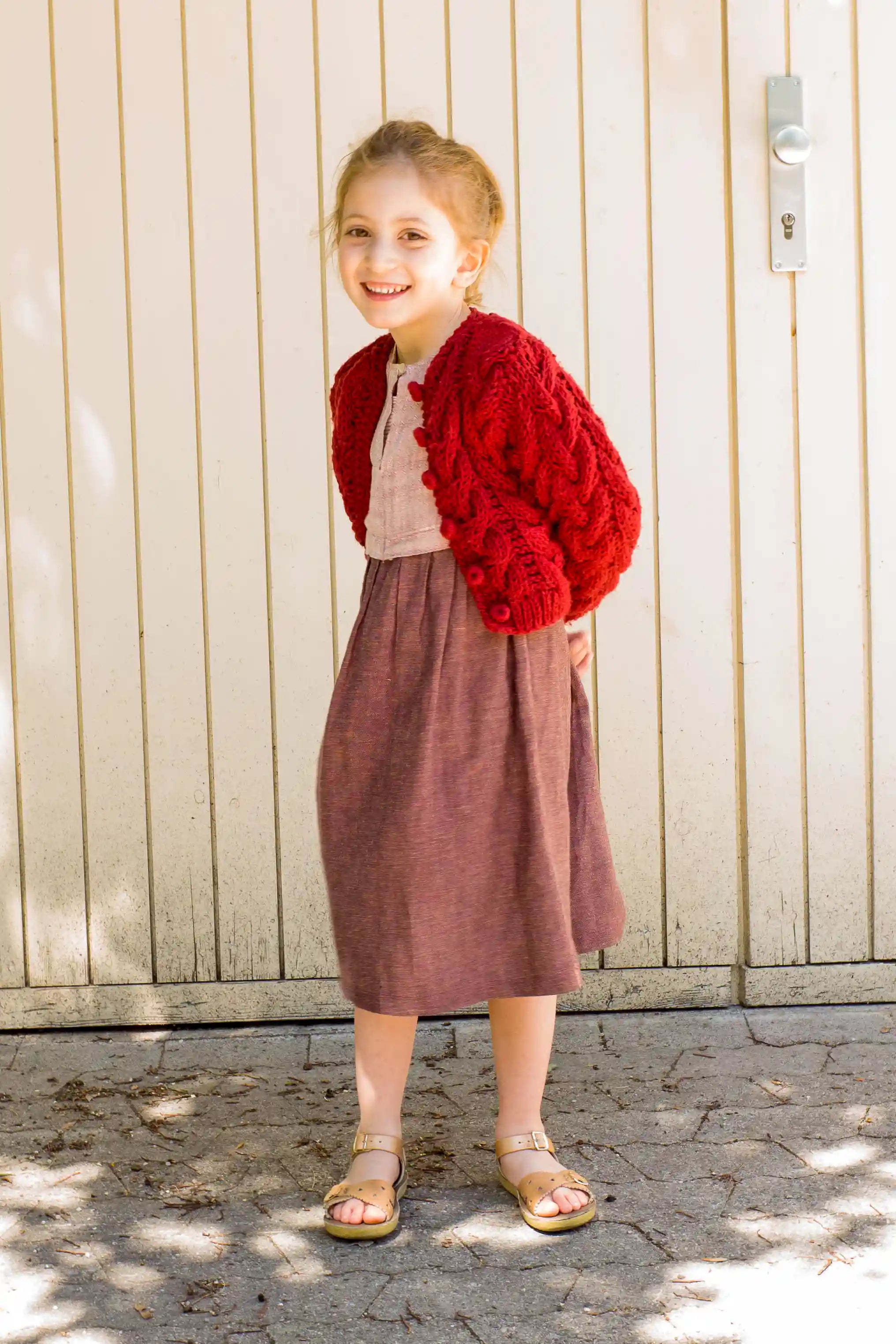 Kinderdirndl in Rosenholz mit kirschroter Strickjacke – Seitenansicht lachend vor einem Garagentor, natürlich inszeniert von Zirbenfüxlein