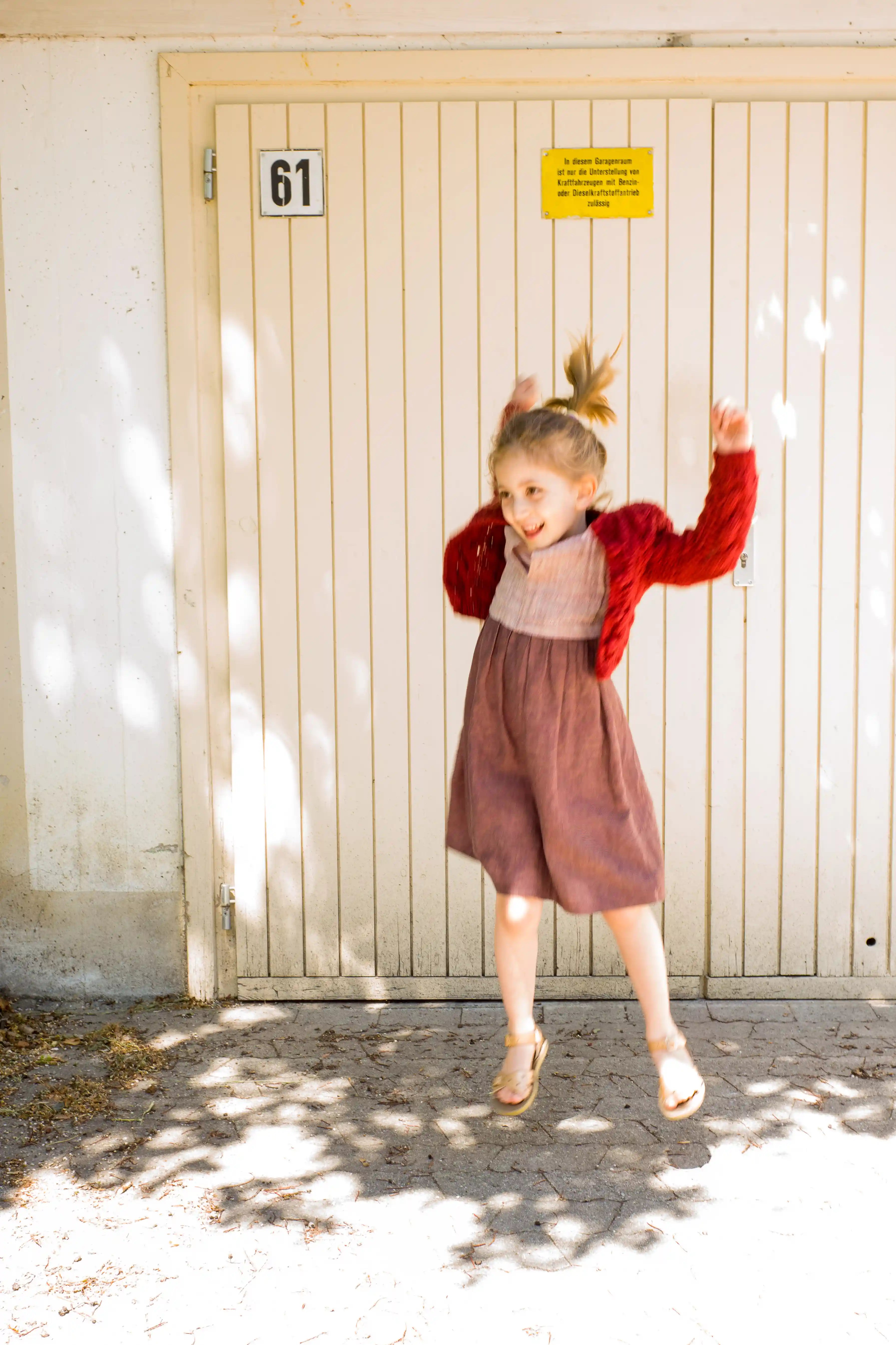 Kinderdirndl in Rosenholz mit kirschroter Strickjacke – fröhlich springend, dynamische Szene mit Unschärfe im Bild, spontan inszeniert von Zirbenfüxlein