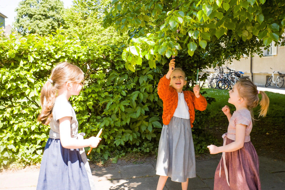Zirbenfüxlein Kinderdirndl in Rosenholz, Tintenblau und Saphir mit Strickjacke, drei Kinder in urbaner Hinterhofszene unter einem Blätterdach, gemeinsam etwas im Baum entdeckend dargestellt, aufmerksam inszeniert von Zirbenfüxlein.