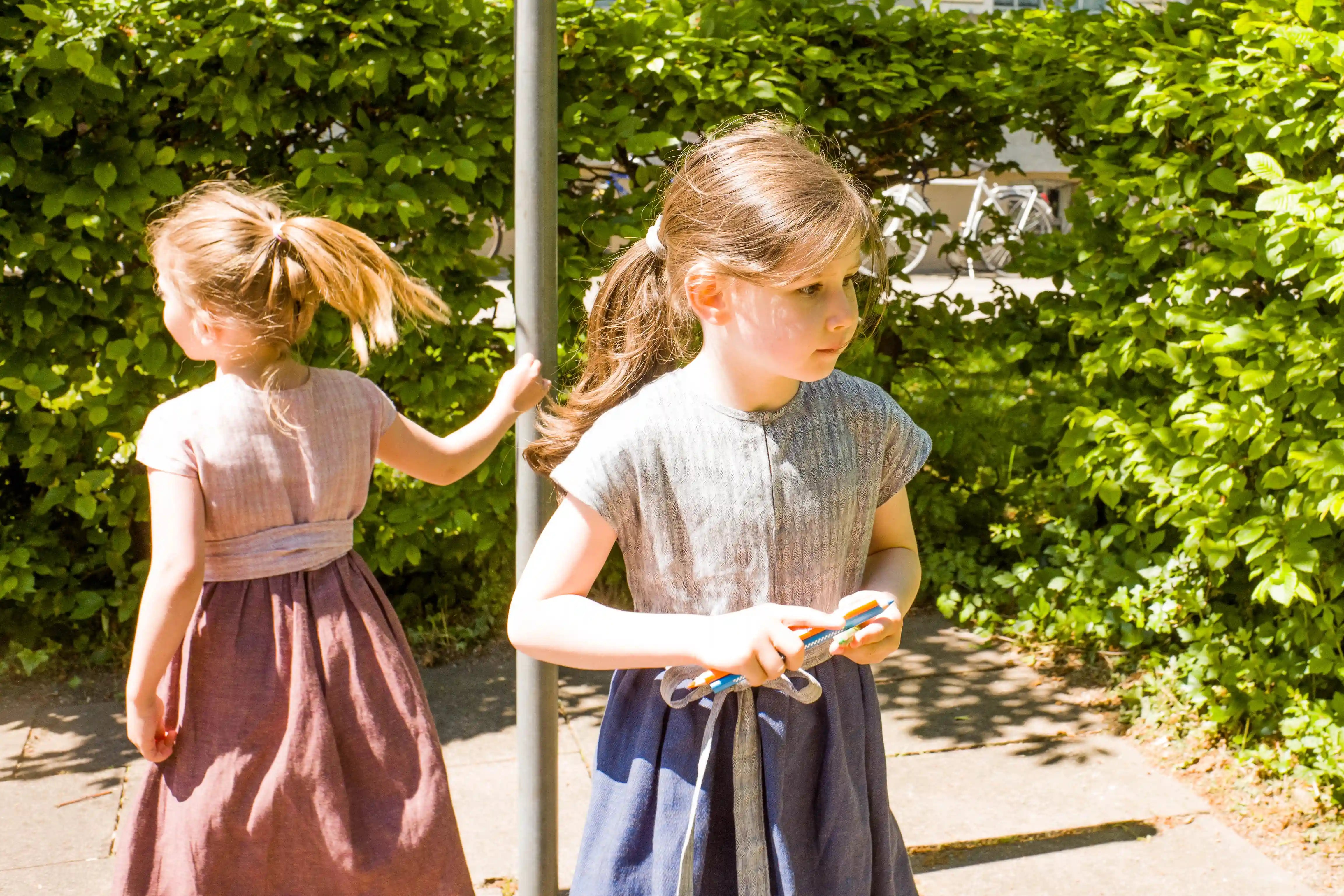 Zirbenfüxlein Kinderdirndl in Rosenholz und Tintenblau mit Schürzenband, zwei Kinder in urbaner Hinterhofszene, eines in Frontansicht und eines in Rückansicht suchend dargestellt, sensibel interpretiert von Zirbenfüxlein.