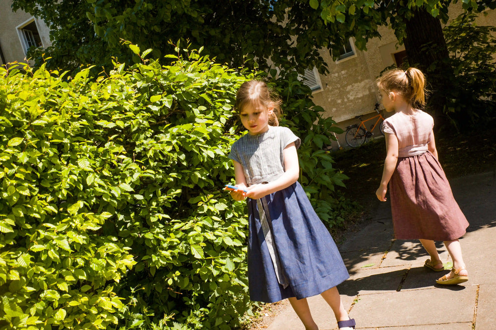 Zirbenfüxlein Kinderdirndl in Rosenholz und Tintenblau mit Schürzenband, zwei Kinder in urbaner Hinterhofszene, eines in Frontansicht und eines in Rückansicht suchend dargestellt, sensibel interpretiert von Zirbenfüxlein.