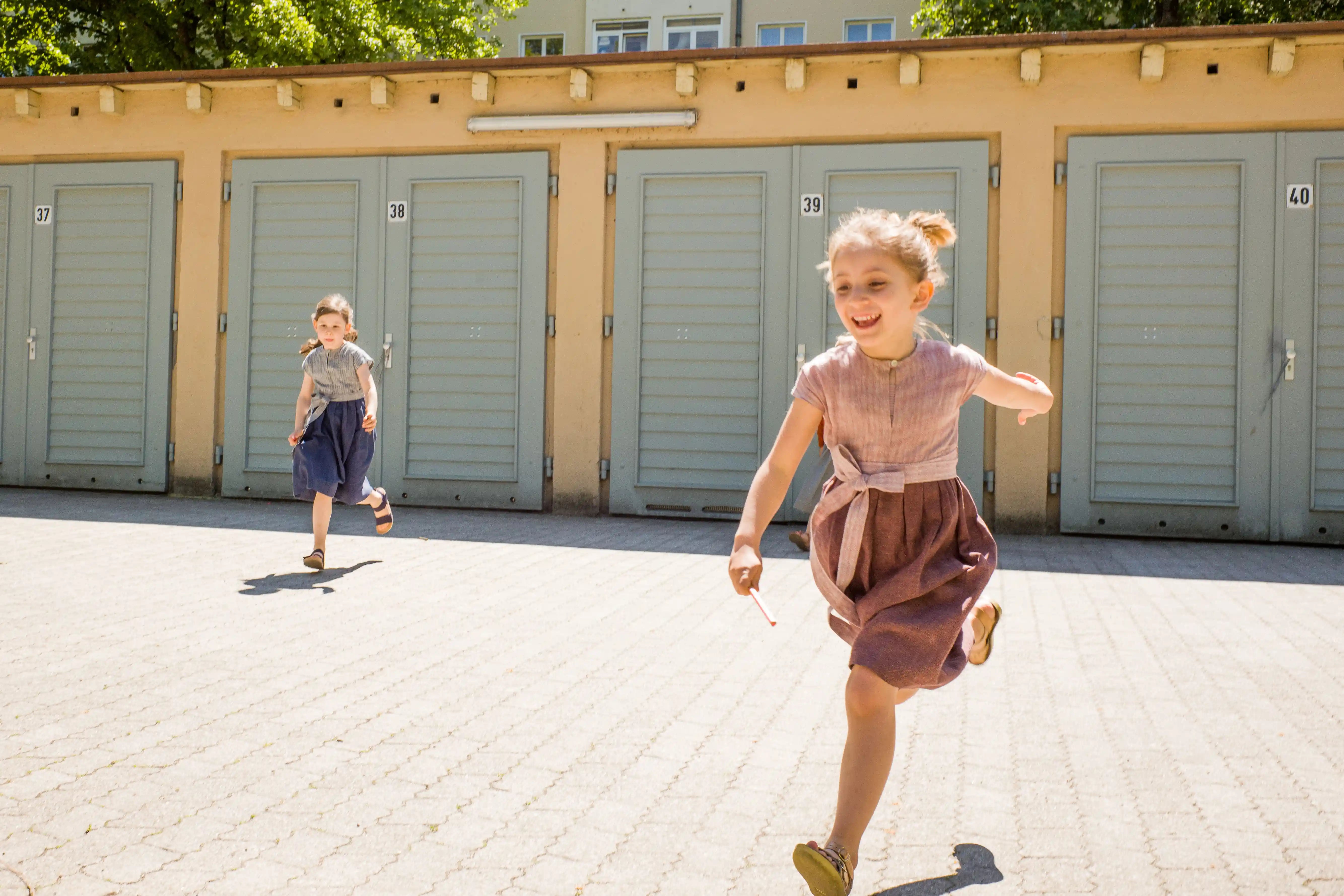 Zirbenfüxlein Kinderdirndl in Rosenholz und Tintenblau mit Schürzenband, zwei Kinder rennen in sonnigem Hinterhof vor Garagentoren, dynamisch eingefangen von Zirbenfüxlein.