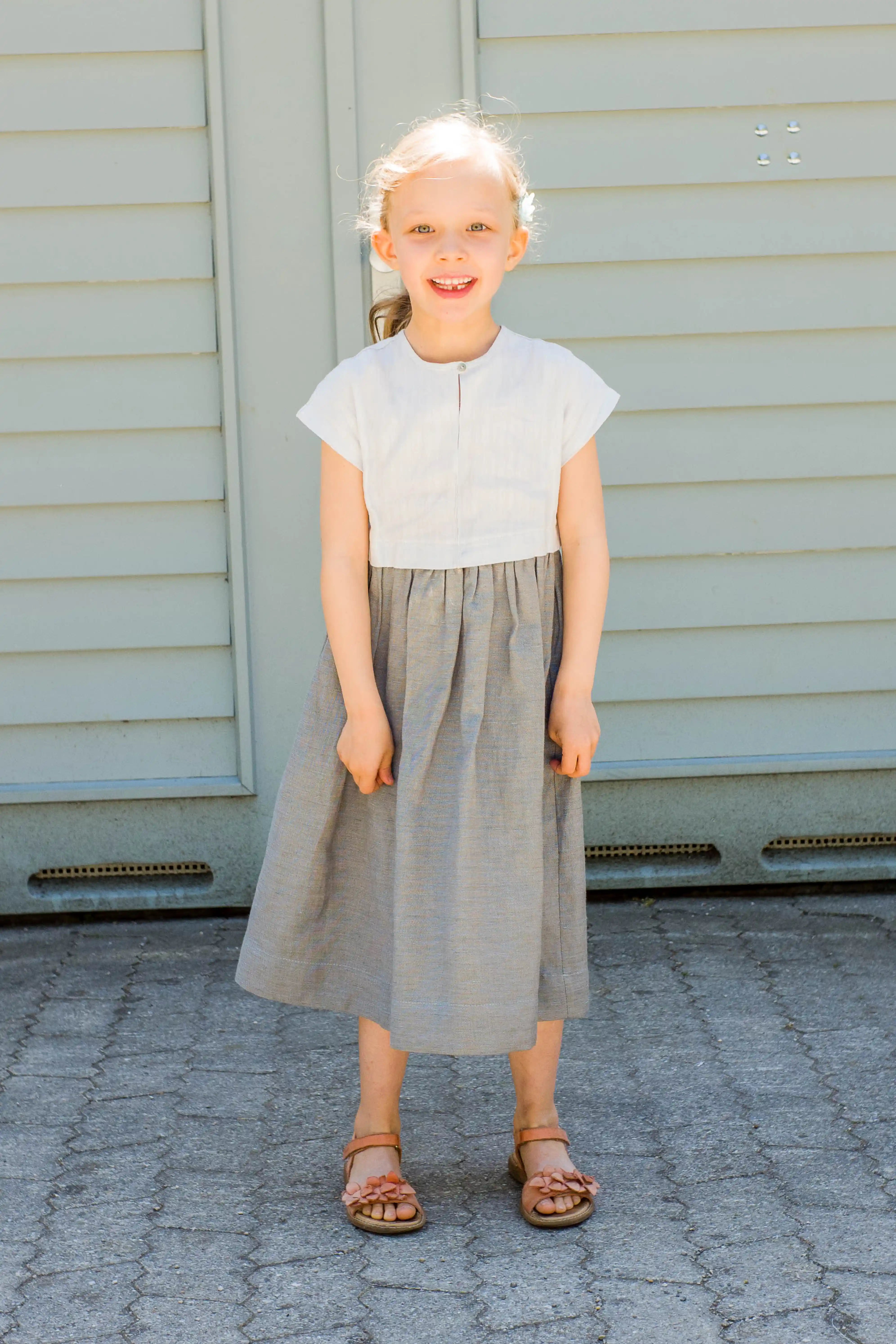 Kinderdirndl in Saphirblau – frontal vor einem Garagentor in heller Sonne stehend, fröhlich und modern inszeniert von Zirbenfüxlein