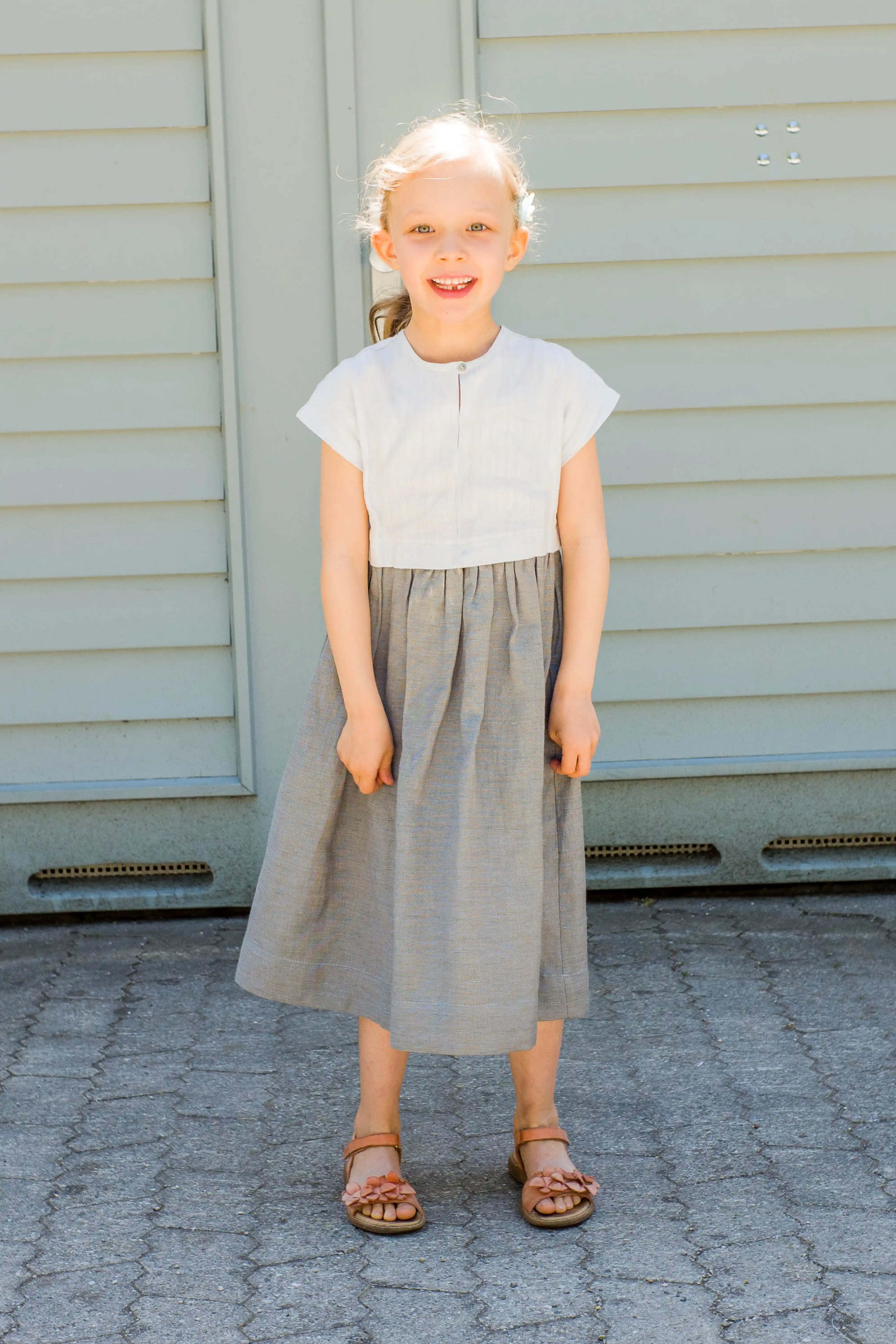 Kinderdirndl in Saphirblau – frontal vor einem Garagentor in heller Sonne stehend, fröhlich und modern inszeniert von Zirbenfüxlein