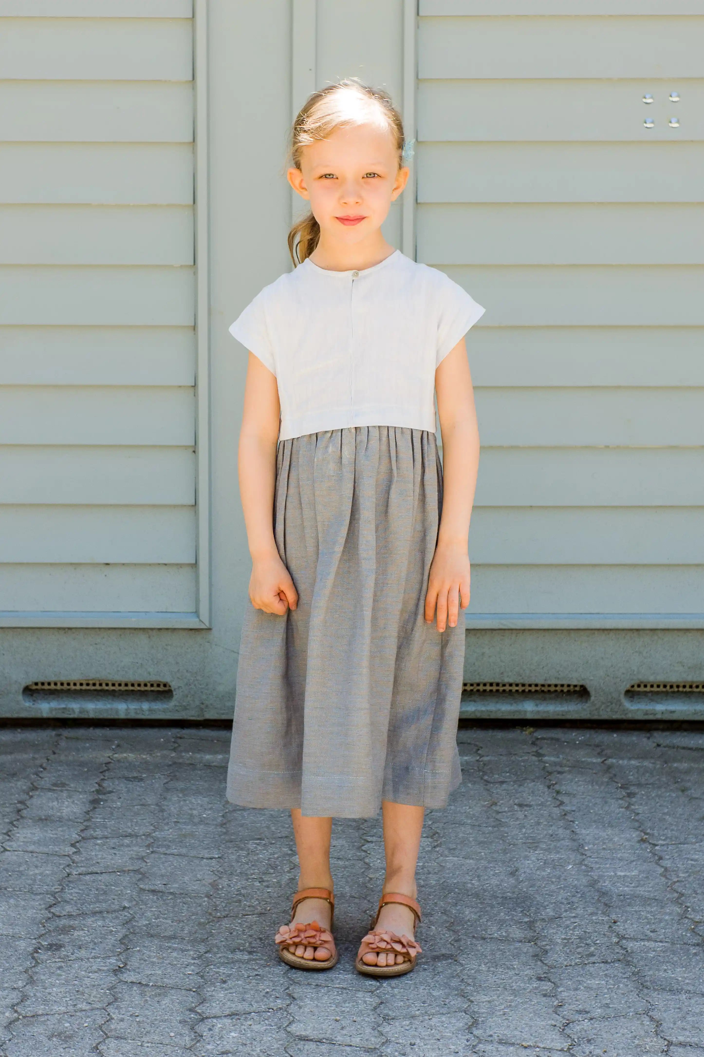Zirbenfüxlein Kinderdirndl Saphir, Frontansicht eines stehenden Kindes vor einem Hinterhof-Garagentor, reduziert dargestellt von Zirbenfüxlein.
