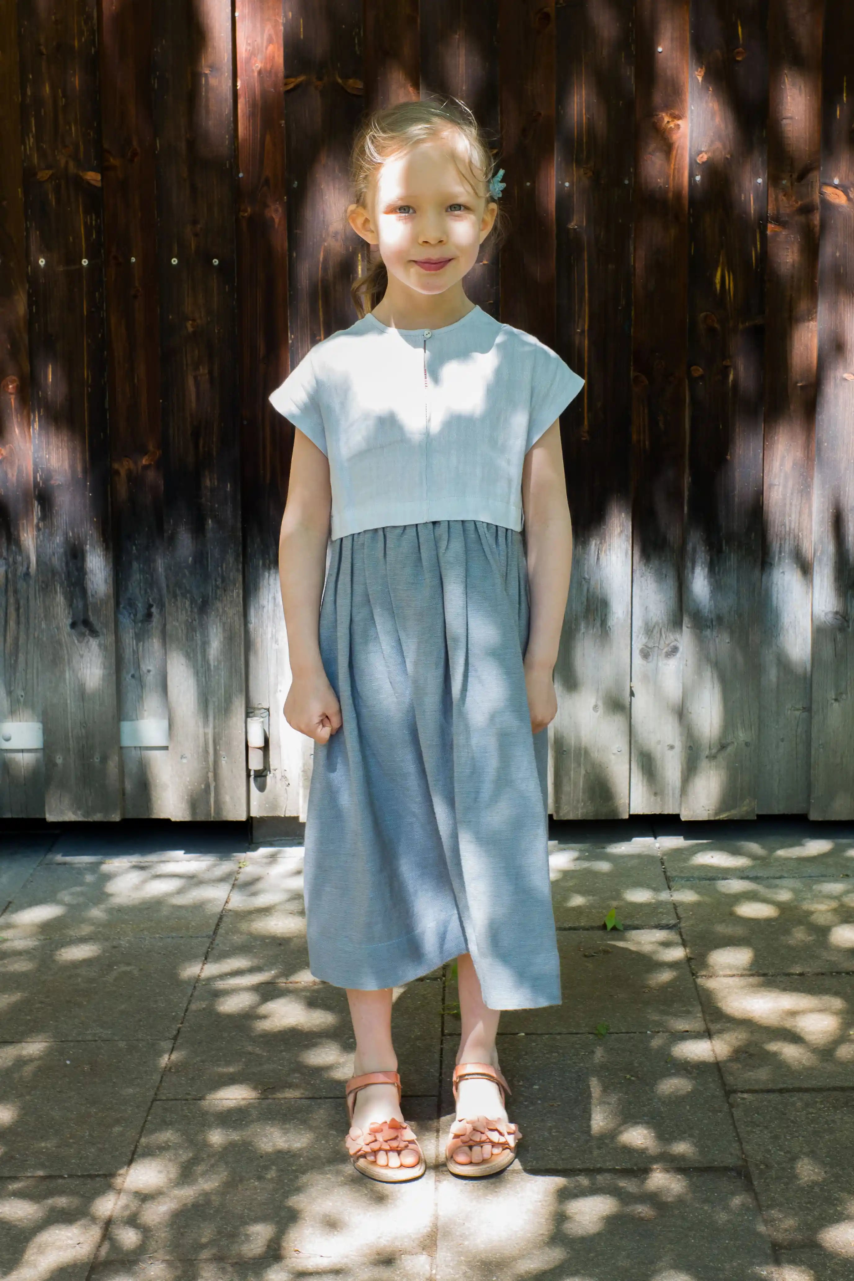 Kinderdirndl in Saphirblau – frontal vor einer Altholzwand stehend im urbanen Setting, markantes Licht- und Schattenspiel, stilvoll inszeniert von Zirbenfüxlein