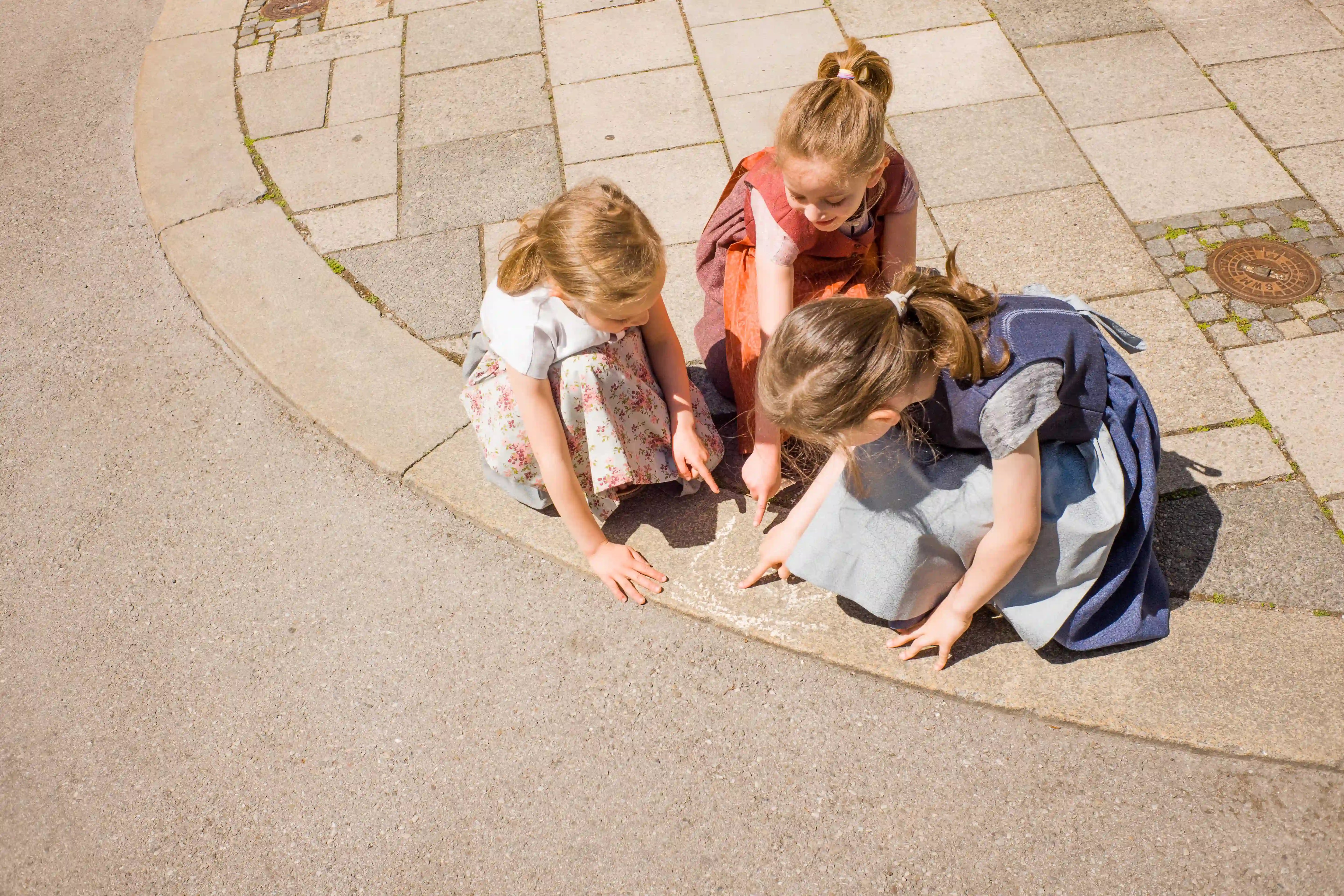 Zirbenfüxlein Kinderdirndl in Saphir, pinke Beere und Tintenblau, drei Kinder in der Hocke an einer sonnigen Straßenecke, gemeinsam eine Kreidemarkierung inspizierend, aufmerksam umgesetzt von Zirbenfüxlein.