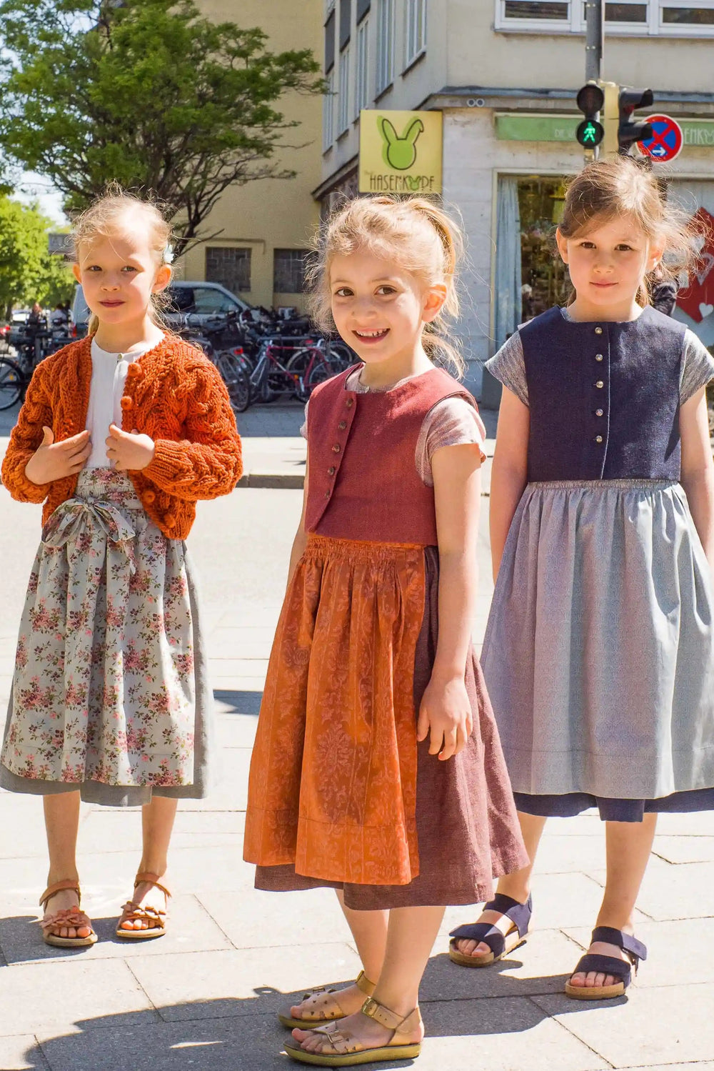 Zirbenfüxlein Kinderdirndl in Saphir, pinke Beere und Tintenblau, drei Kinder an einer belebten Kreuzung beieinander stehend in urbaner Szene, harmonisch inszeniert von Zirbenfüxlein.