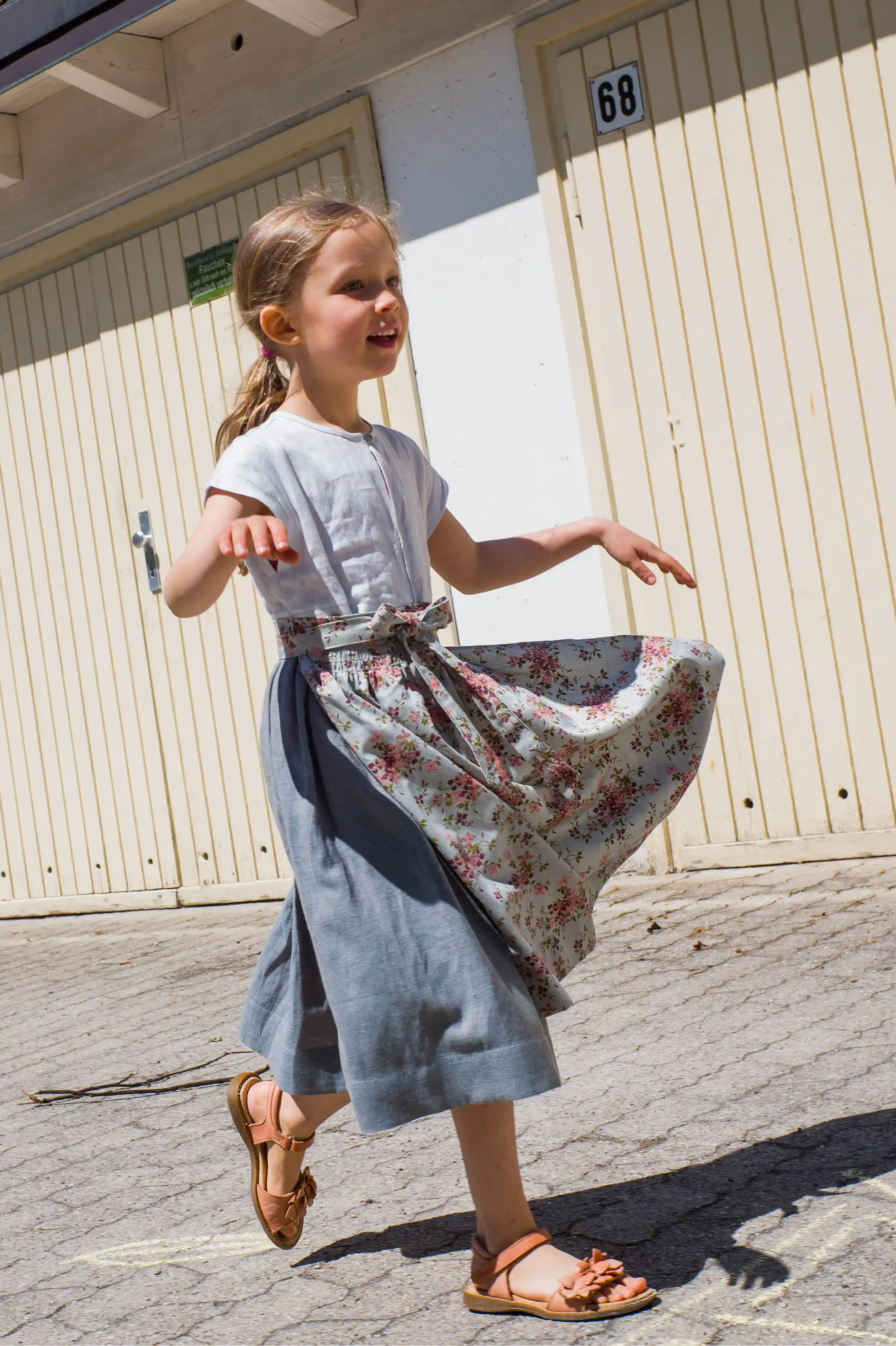 Zirbenfüxlein Kinderdirndl Saphir aus Leinen mit hellblauer Schürze mit Blumenmuster, Kind läuft entlang einer Garageneinfahrt, Schürze in Bewegung vor urbanen Garagentoren, lebendig umgesetzt von Zirbenfüxlein.