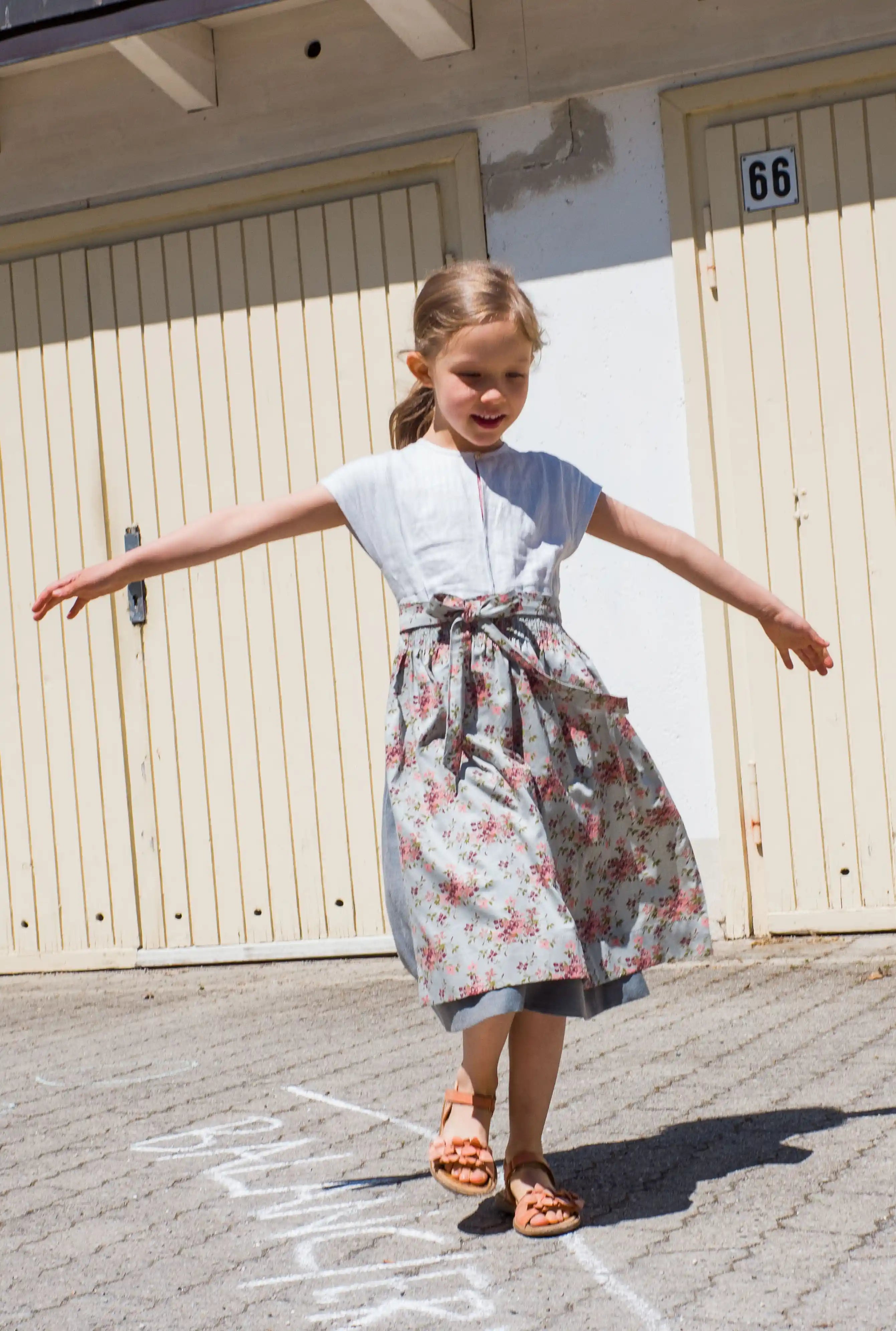 Zirbenfüxlein Kinderdirndl Saphir aus Leinen mit hellblauer Schürze mit Blumenmuster, Frontansicht eines balancierenden Kindes auf Kreidelinie vor urbanen Garagentoren, zeitgemäß inszeniert von Zirbenfüxlein.
