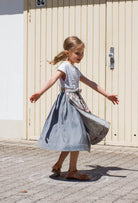 Zirbenfüxlein Kinderdirndl Saphir aus Leinen mit hellblauer Schürze mit Blumenmuster, schwungvolle Drehung mit ausgestreckten Armen in Frontansicht vor urbanen Garagentoren, lebendig umgesetzt von Zirbenfüxlein.